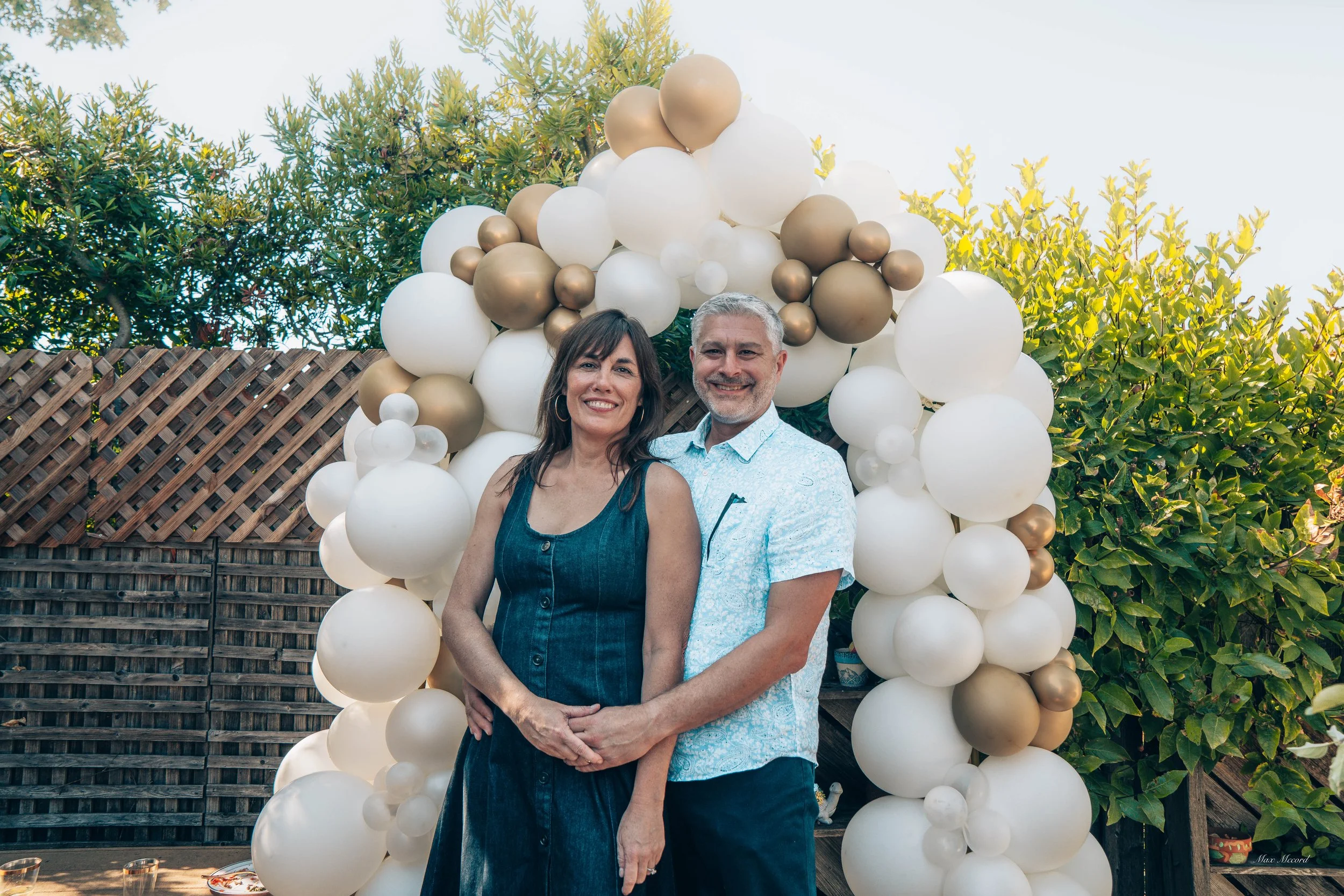A smiling couple stands together outdoors in front of a balloon arch decorated with white and gold balloons, with greenery and trees in the background.