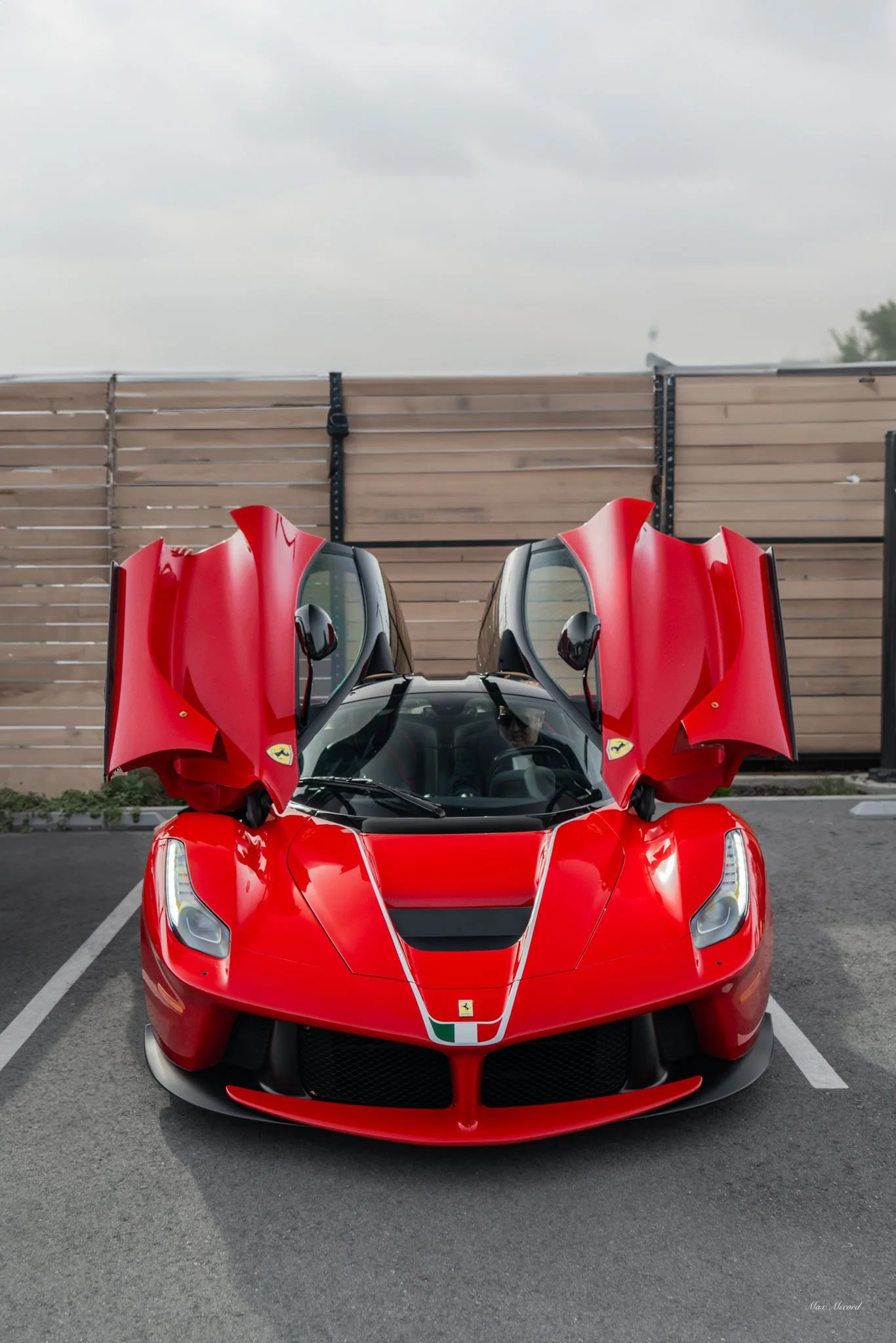 Red Ferrari sports car with gull-wing doors open, parked in an outdoor lot against a wooden fence and overcast sky.