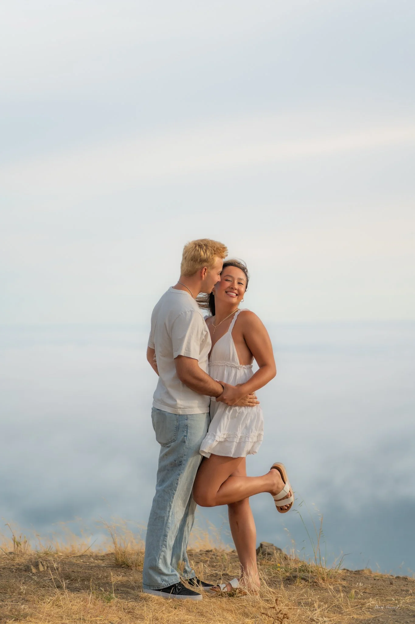 A couple embracing on a grassy hilltop with a cloudy sky in the background. The woman is wearing a white dress and sandals, and is smiling as she leans into the man. The man is dressed in a light-colored t-shirt and jeans, holding the woman close.