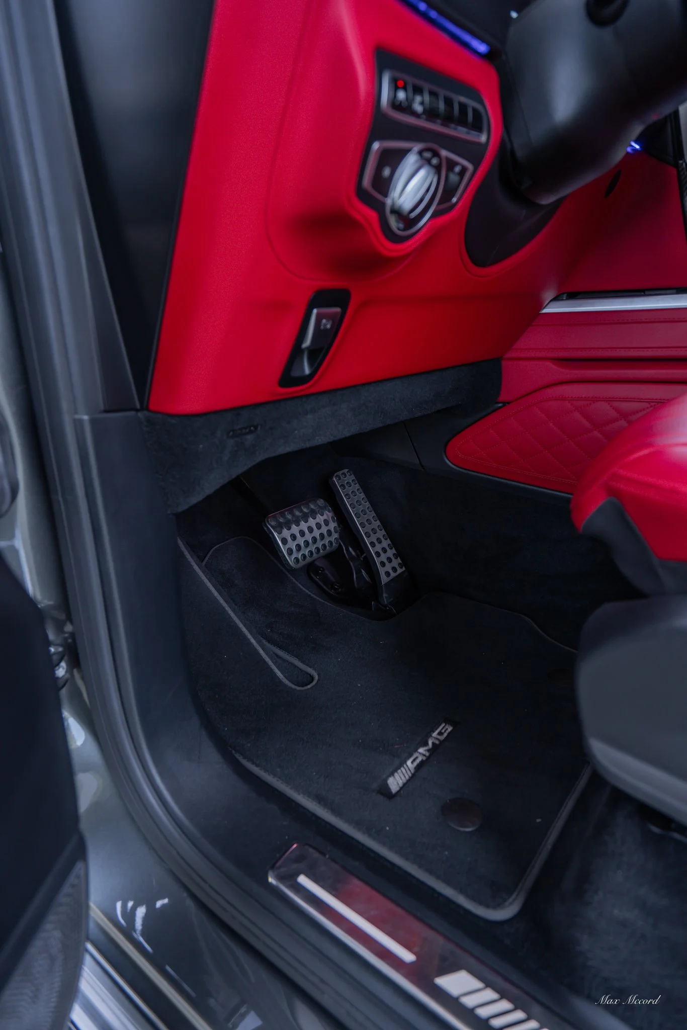 The interior of a luxury car focusing on the driver side, featuring red leather seats, a red dashboard, and a black floor mat with the AMG logo. The car has sporty metal pedals.