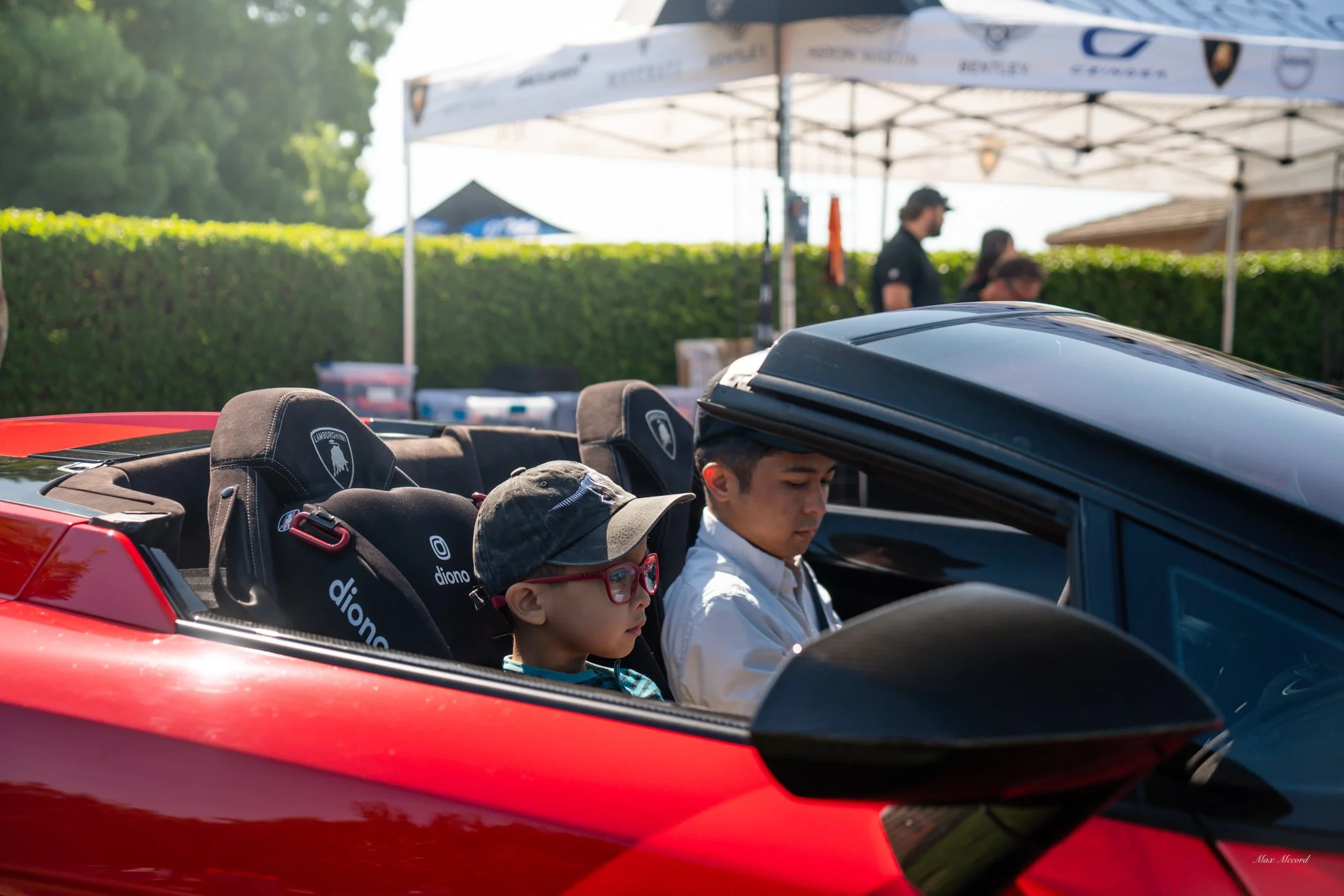 Two children sitting inside a red Lamborghini, with one child wearing glasses and a cap, and the other looking down and focused. The background shows people standing under a canopy and a hedge.