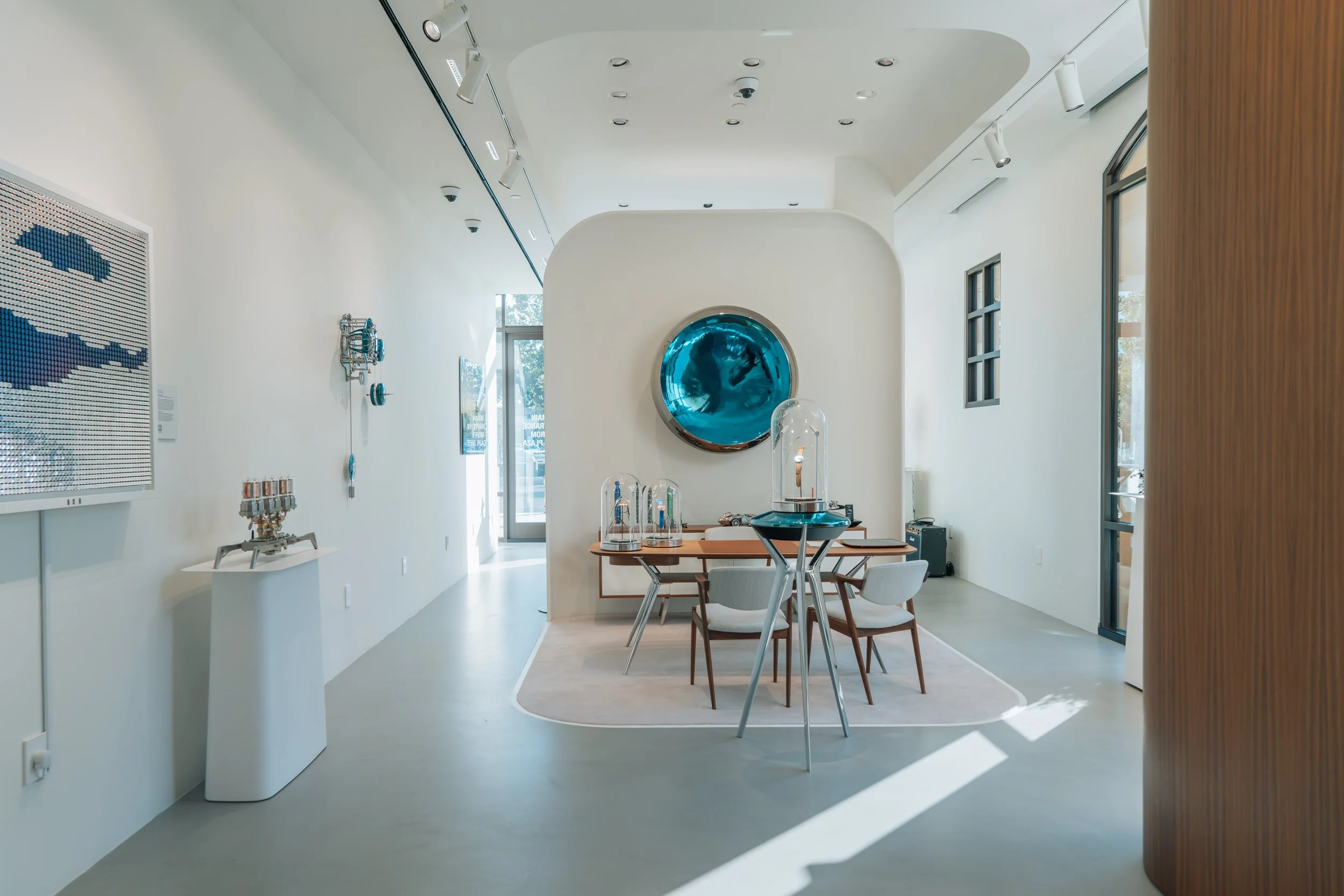 Interior of a modern art gallery featuring a circular blue abstract artwork on a white wall, a glass display case with a sculpture, and various art pieces on the walls, with natural light streaming in through large windows.