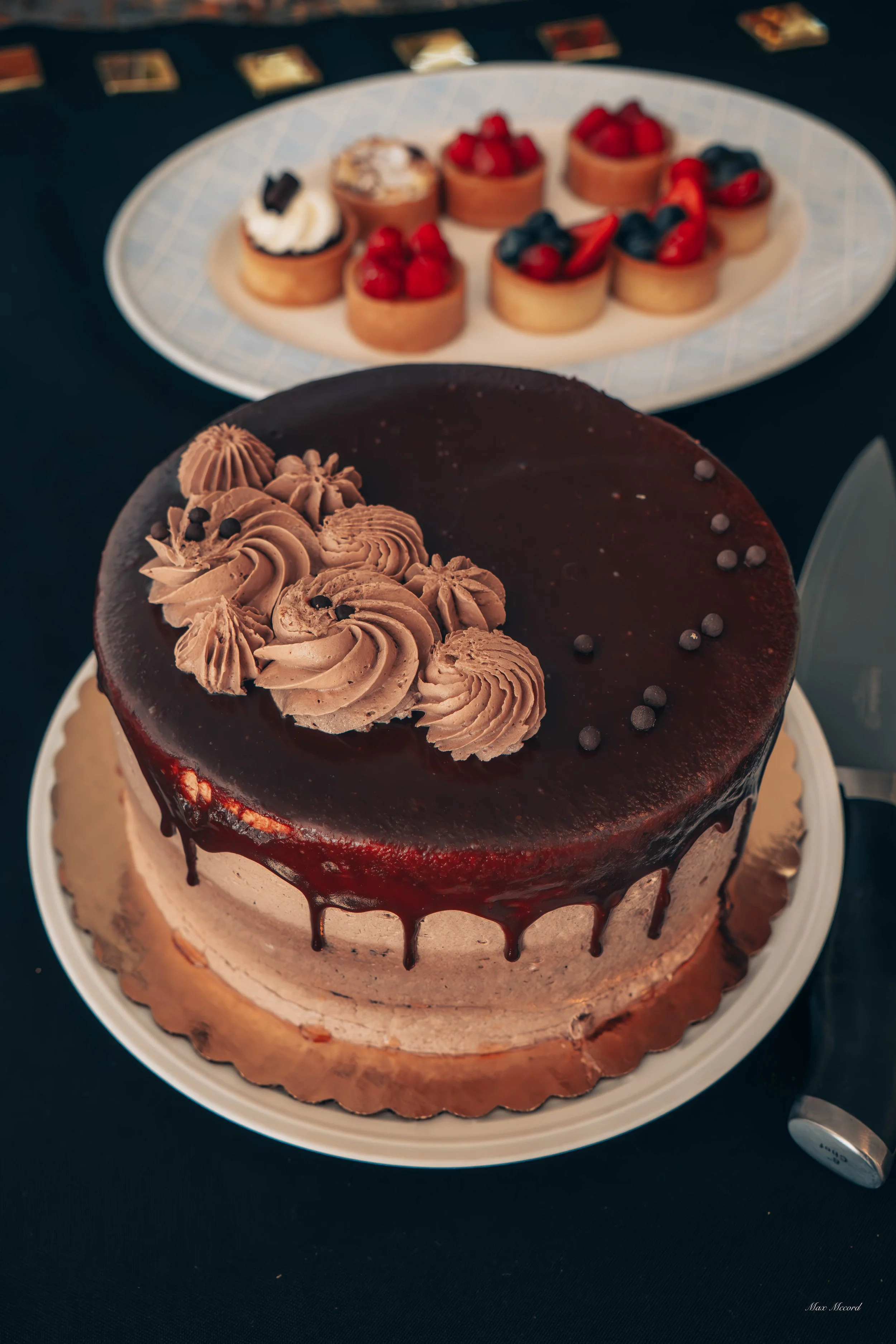 Chocolate cake with chocolate frosting and decorative swirls on top, placed on a white cake board. In the background, a platter of small fruit tarts topped with strawberries, raspberries, and blueberries. The desserts are on a dark table.
