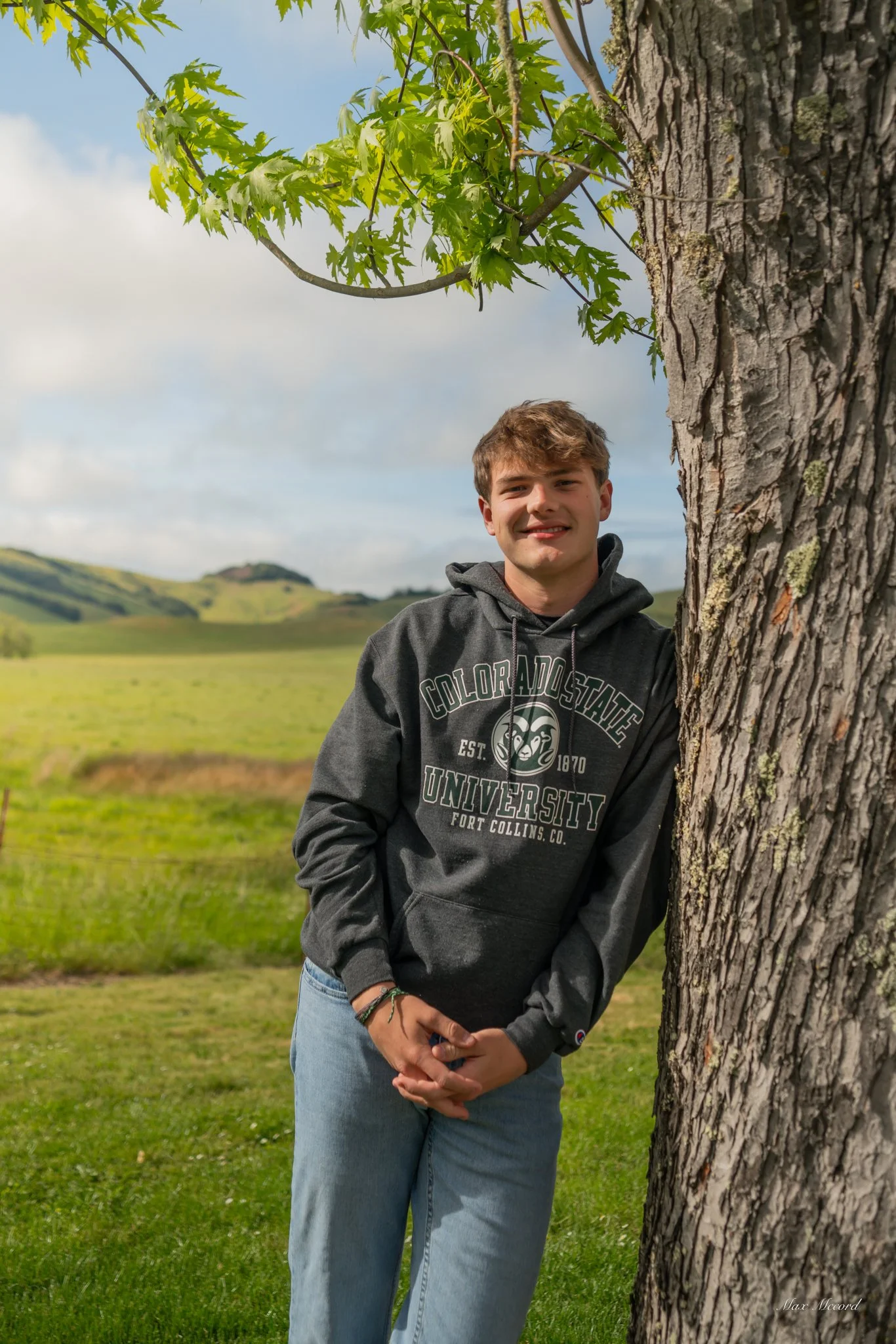 A young man leaning against a tree in a grassy field with rolling hills in the background, wearing a gray hoodie with 'Colorado State University' printed on it, and smiling at the camera.