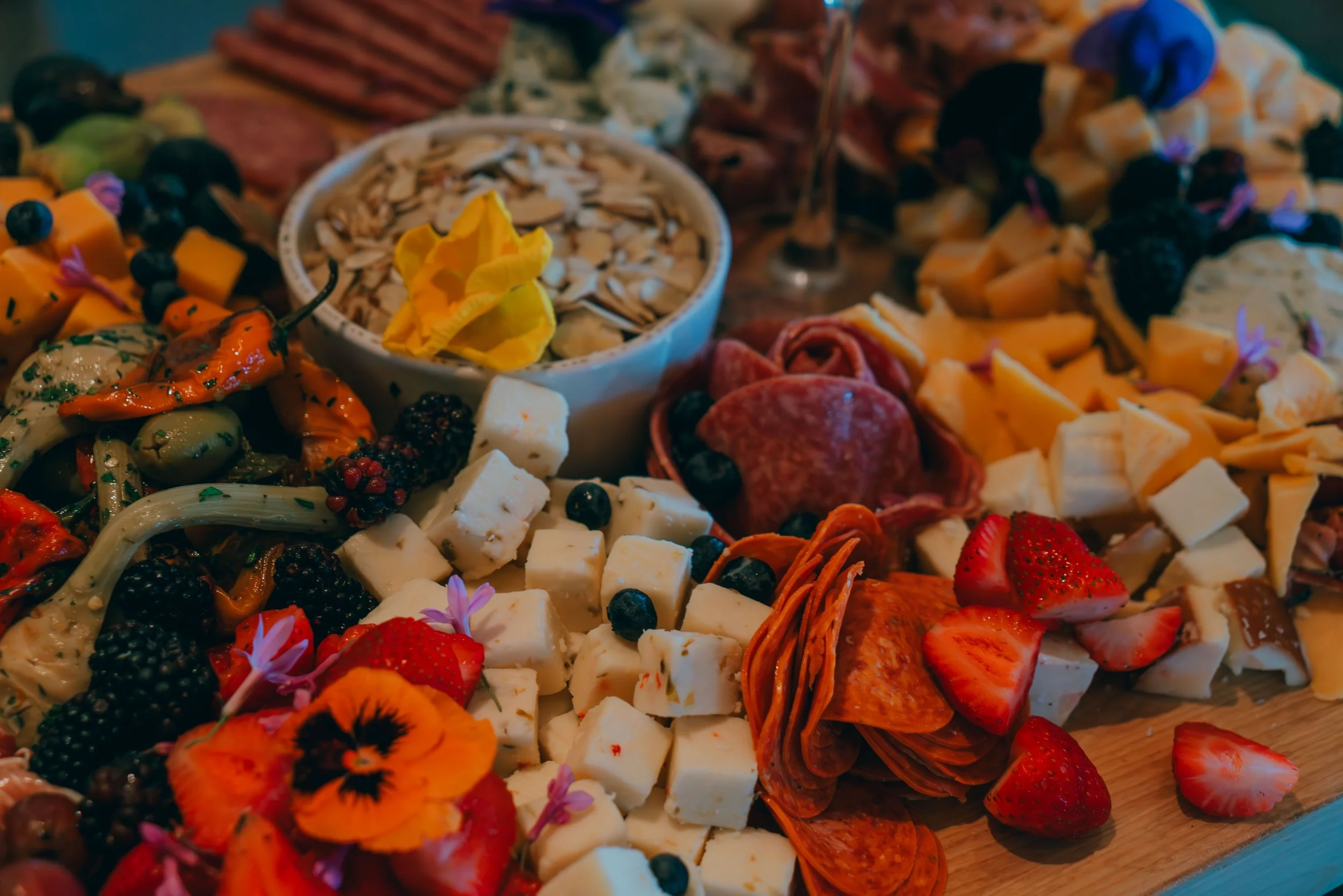 A cheese and fruit platter featuring sliced strawberries, blueberries, blackberries, sliced cheese, cured meats, small cubes of cheese, edible flowers, a bowl of sliced almonds, and various vegetables.