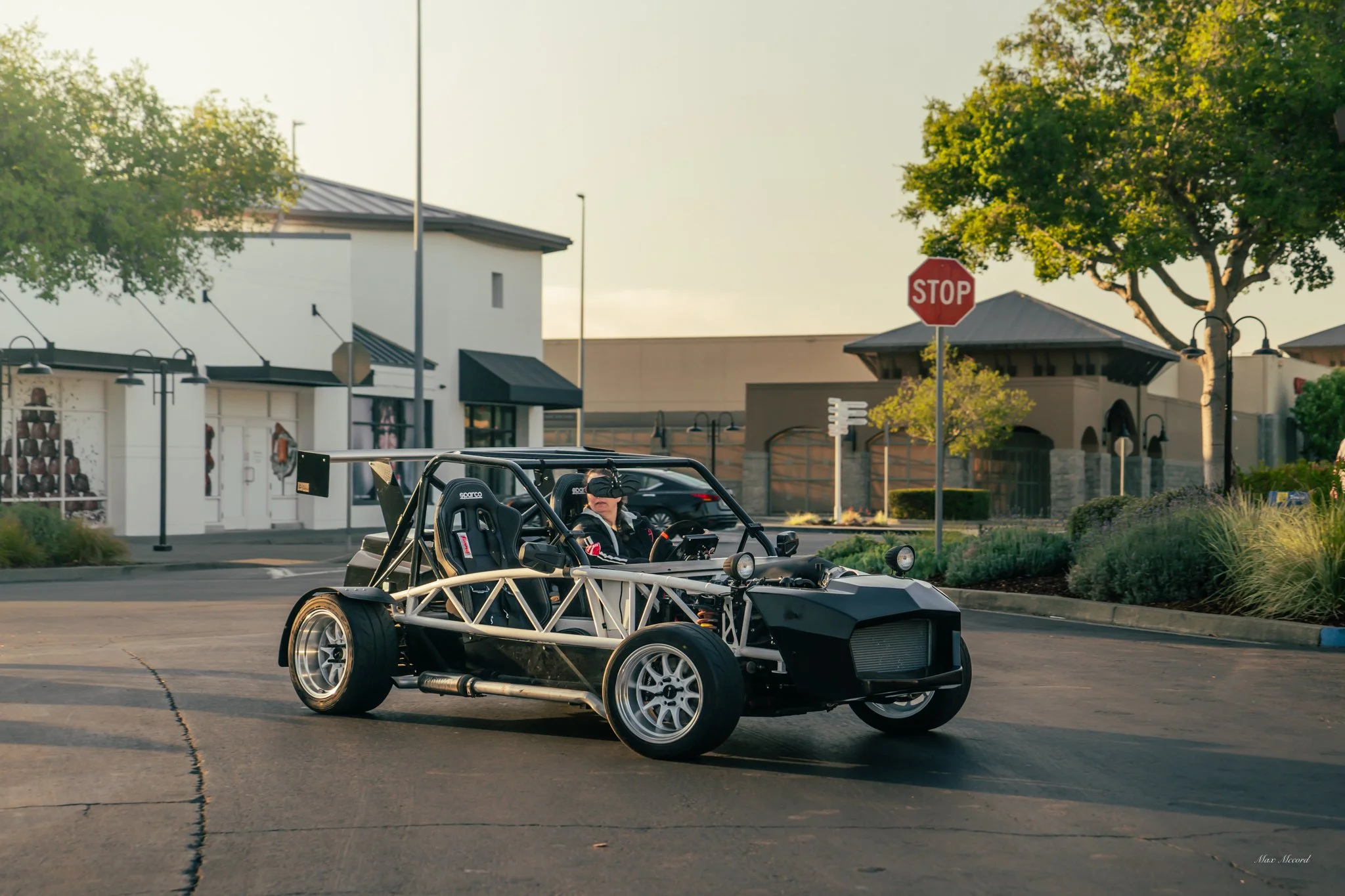 A person driving a vintage-style open-frame race car in a parking lot near stores and trees.