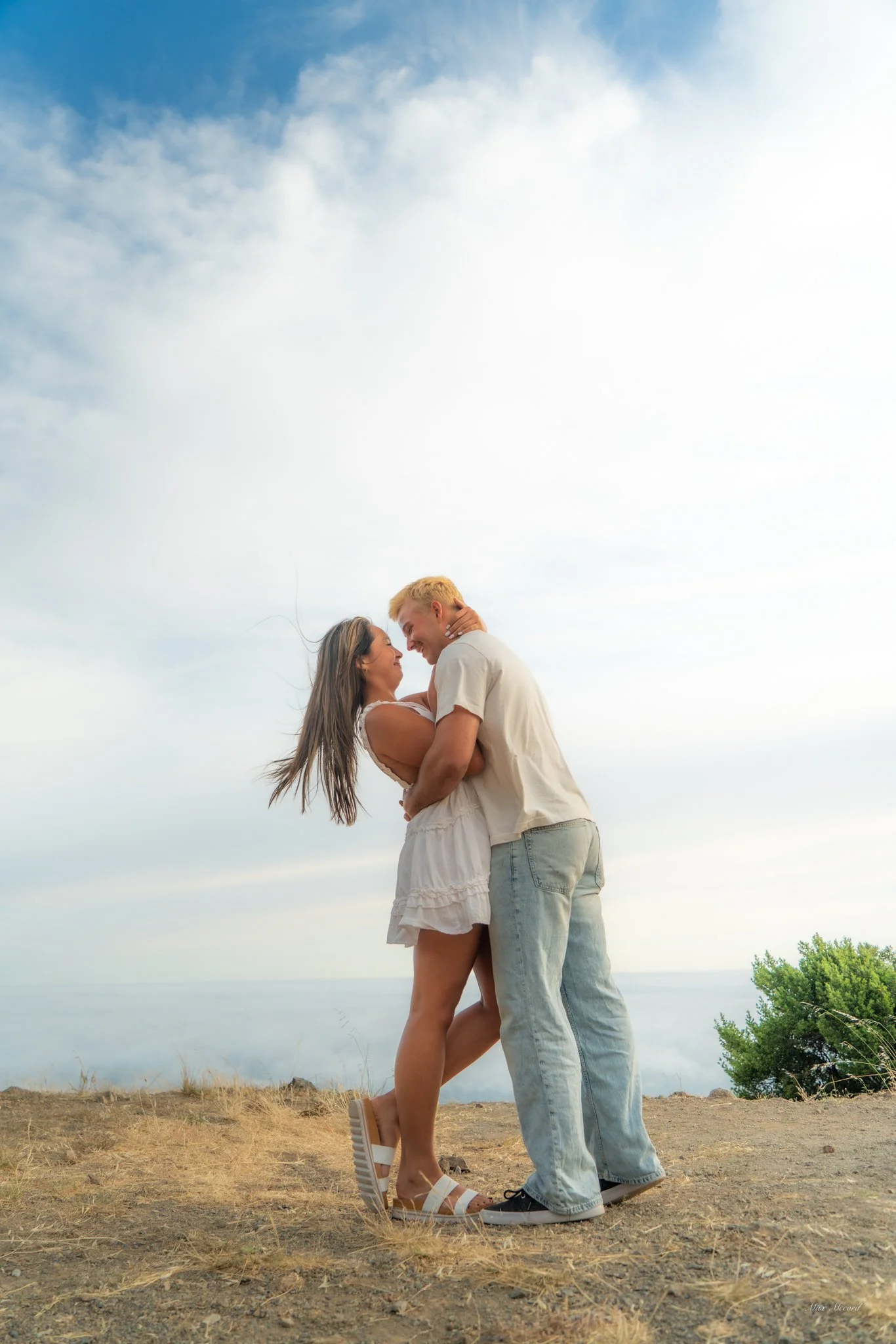 A young couple sharing a joyful embrace outdoors on a clear day, with the woman in a white dress and the man in casual attire, standing on dry grass under a partly cloudy sky.