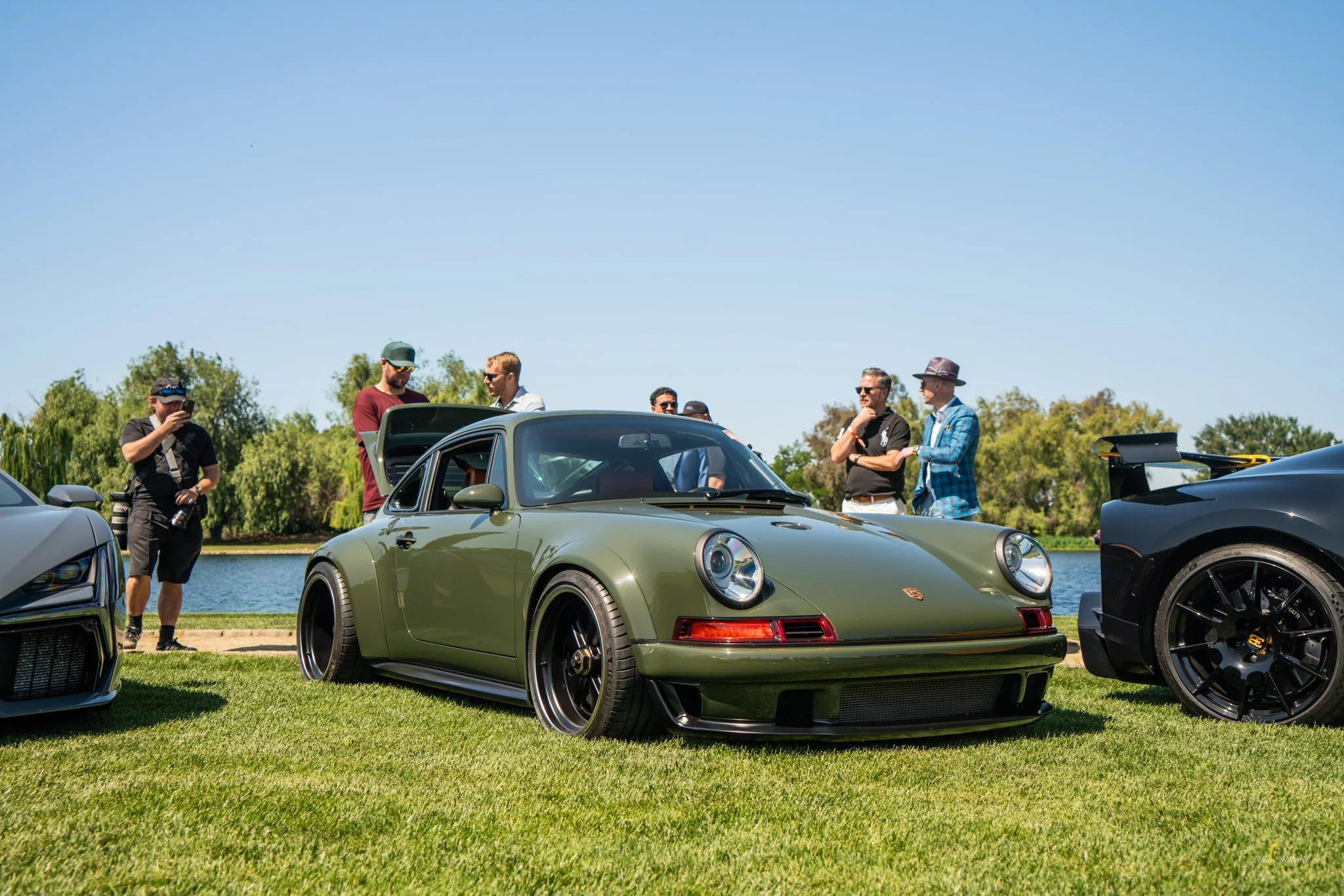 A vintage green Porsche 911 on display at an outdoor car show, with a group of people observing and discussing it near a lake under a clear blue sky.