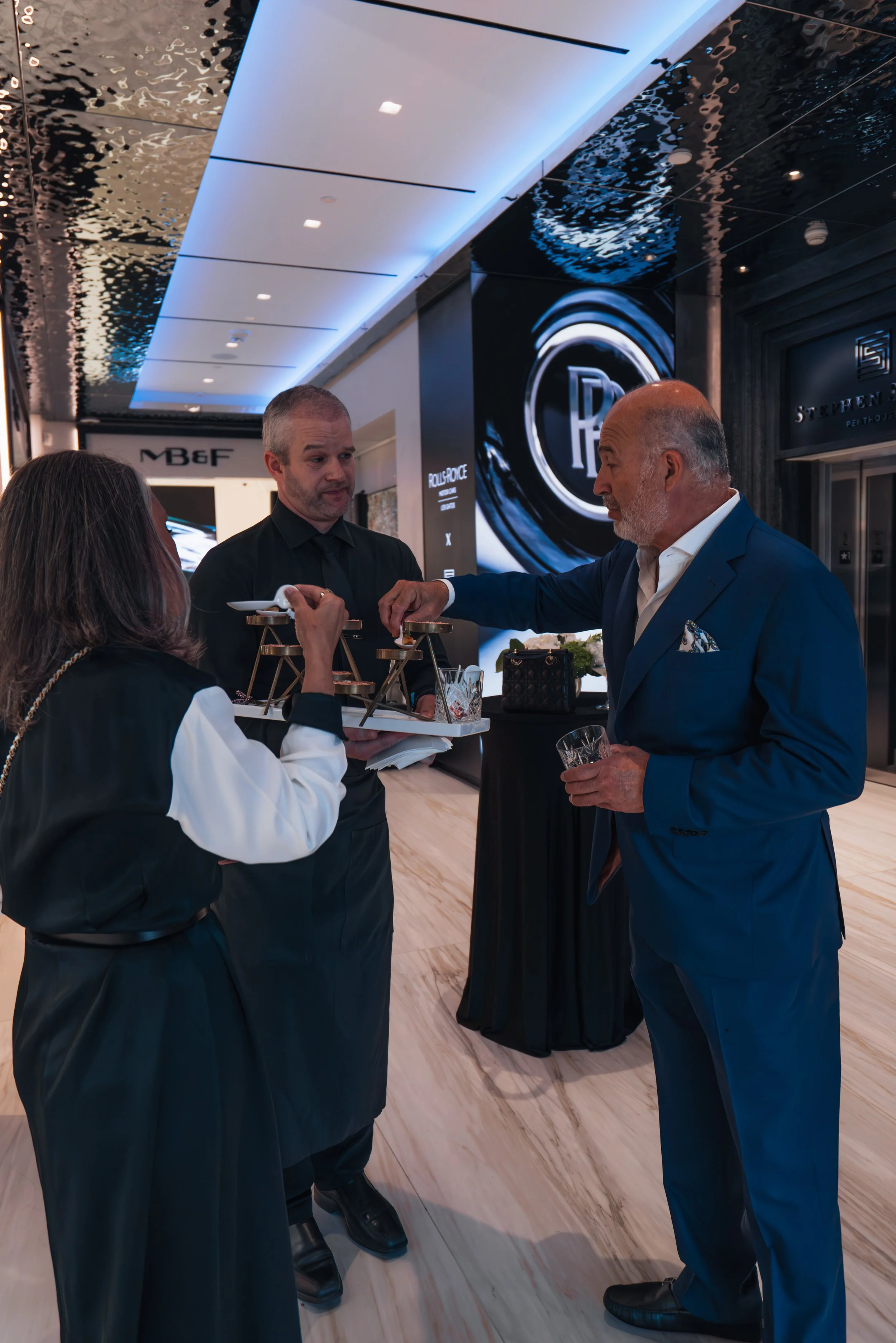 Three people, two men and one woman, are interacting at a social event in a modern, upscale venue. The woman holds a tiered tray of small appetizers or desserts, while one of the men, dressed in a blue suit, is reaching for a treat. The other man in 