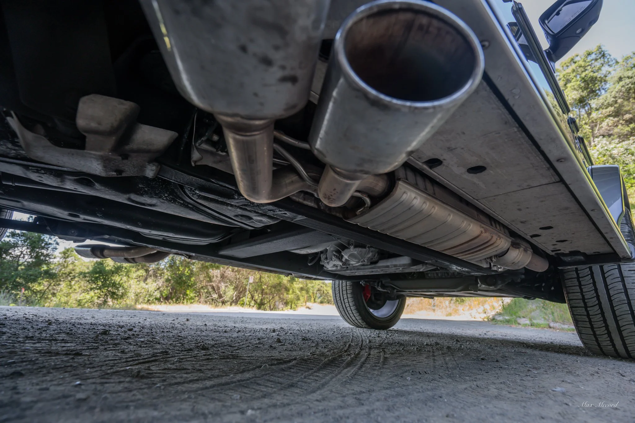 Underneath view of a vehicle showing the exhaust pipes, muffler, and tires on a dirt road with trees in the background.