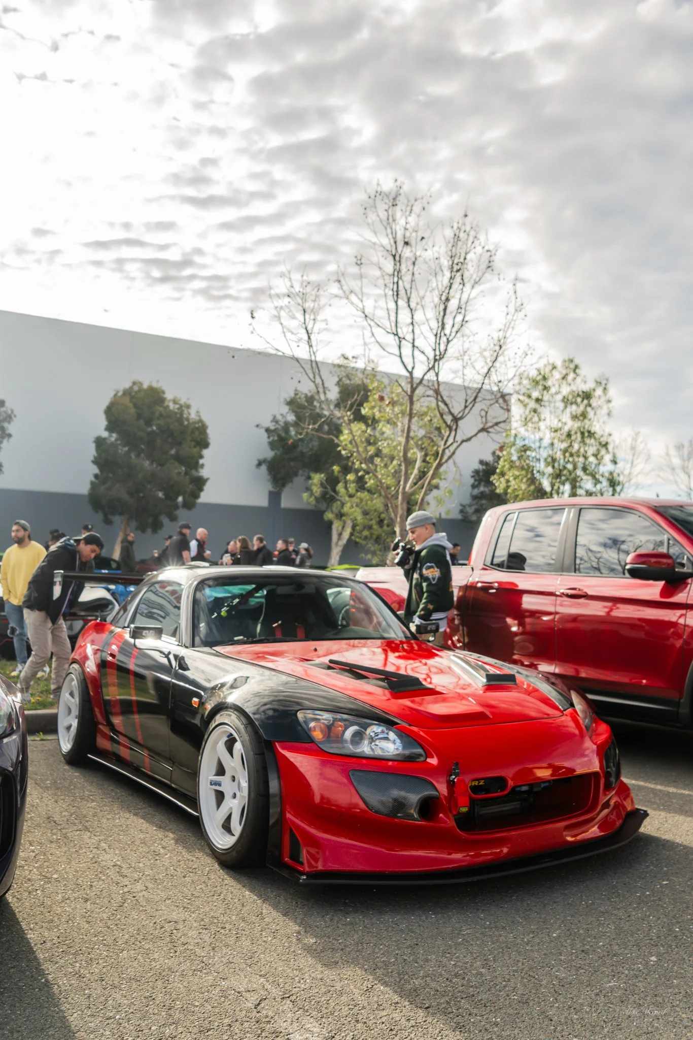 A red and black modified Honda S2000 sports car with white wheels parked in a parking lot during an outdoor car event, with people in the background.