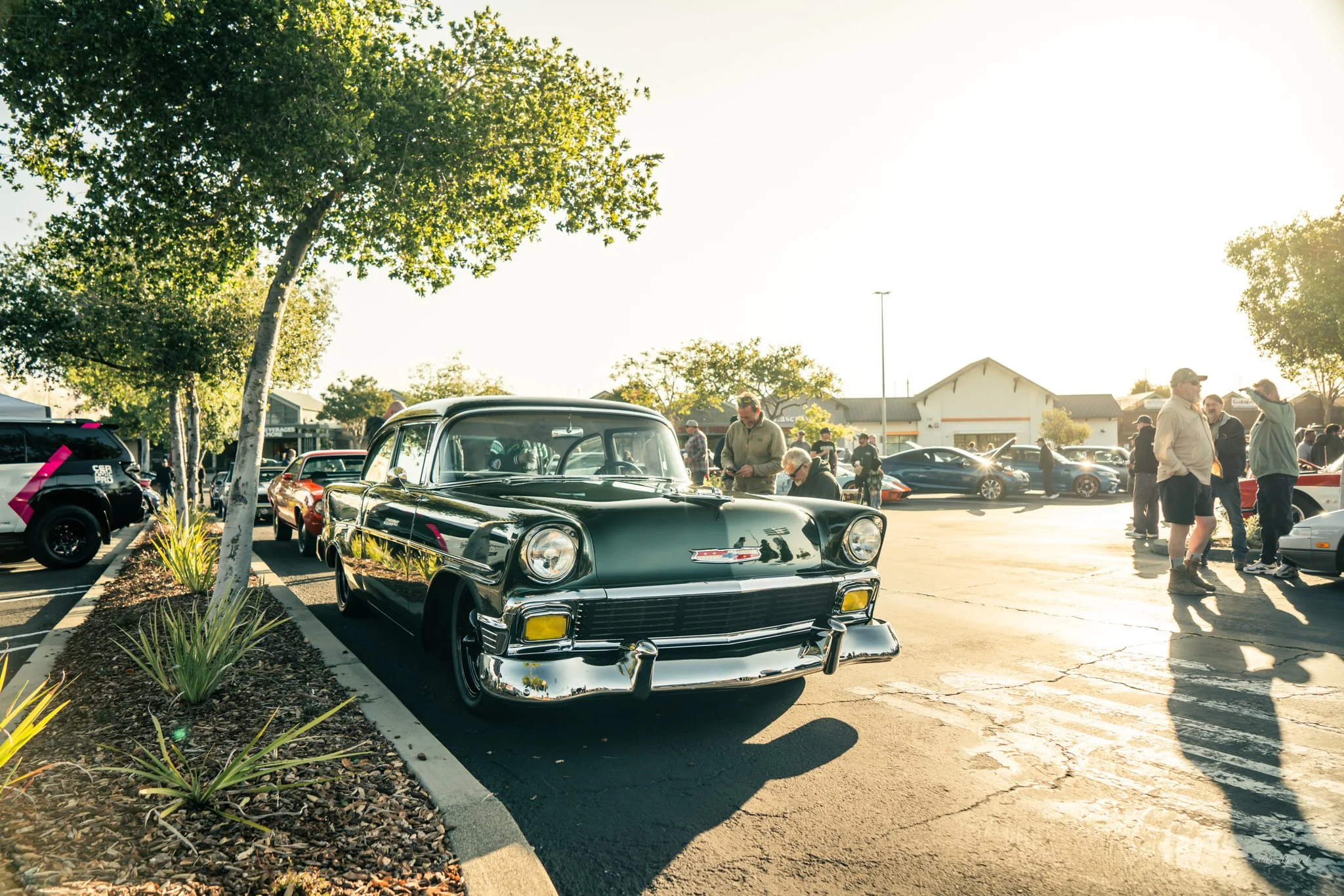 A parking lot filled with vintage and modern cars, people walking and chatting, with trees and a building in the background, illuminated by sunlight.