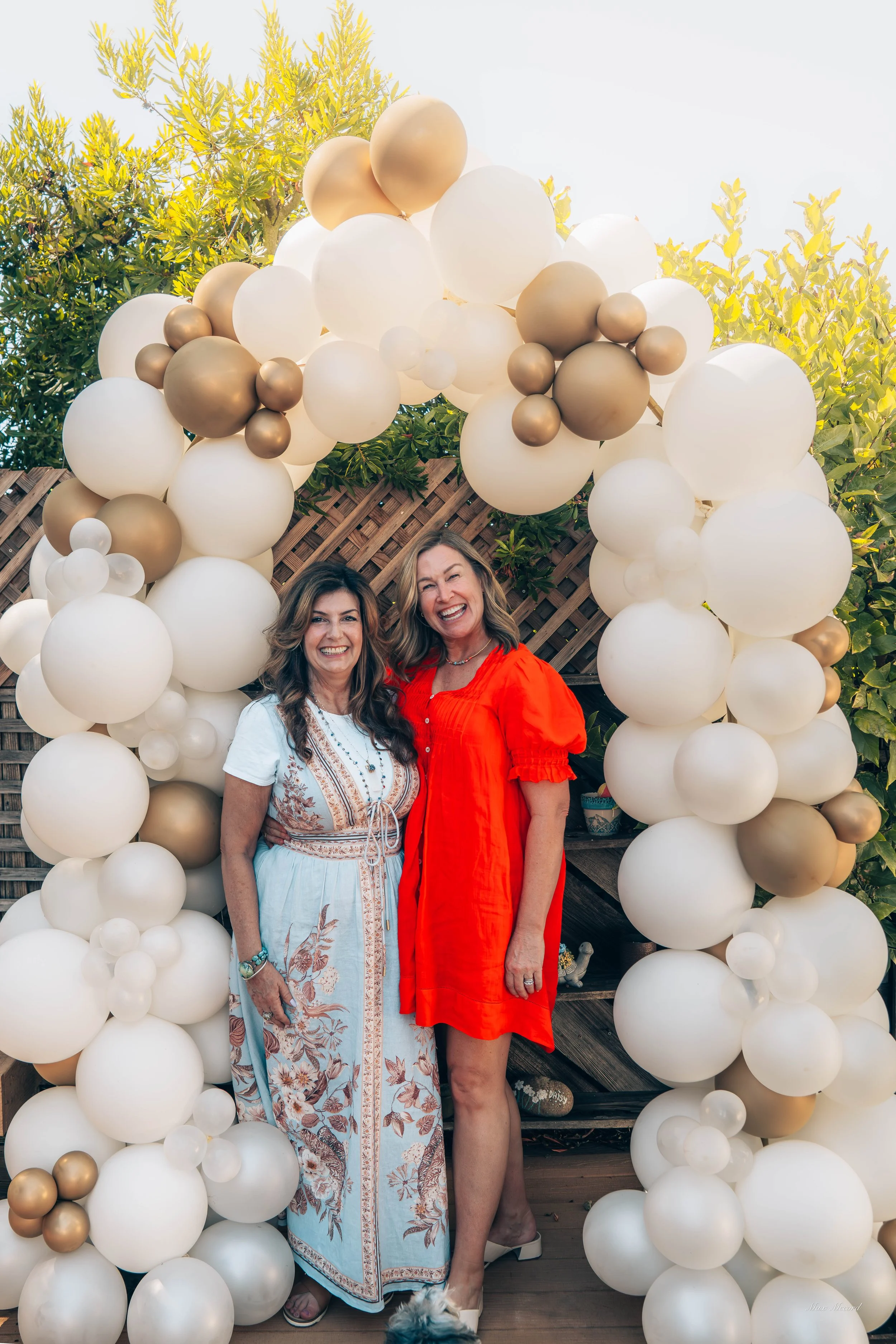 Two women smiling and standing under an arch made of white, gold, and beige balloons outdoors.