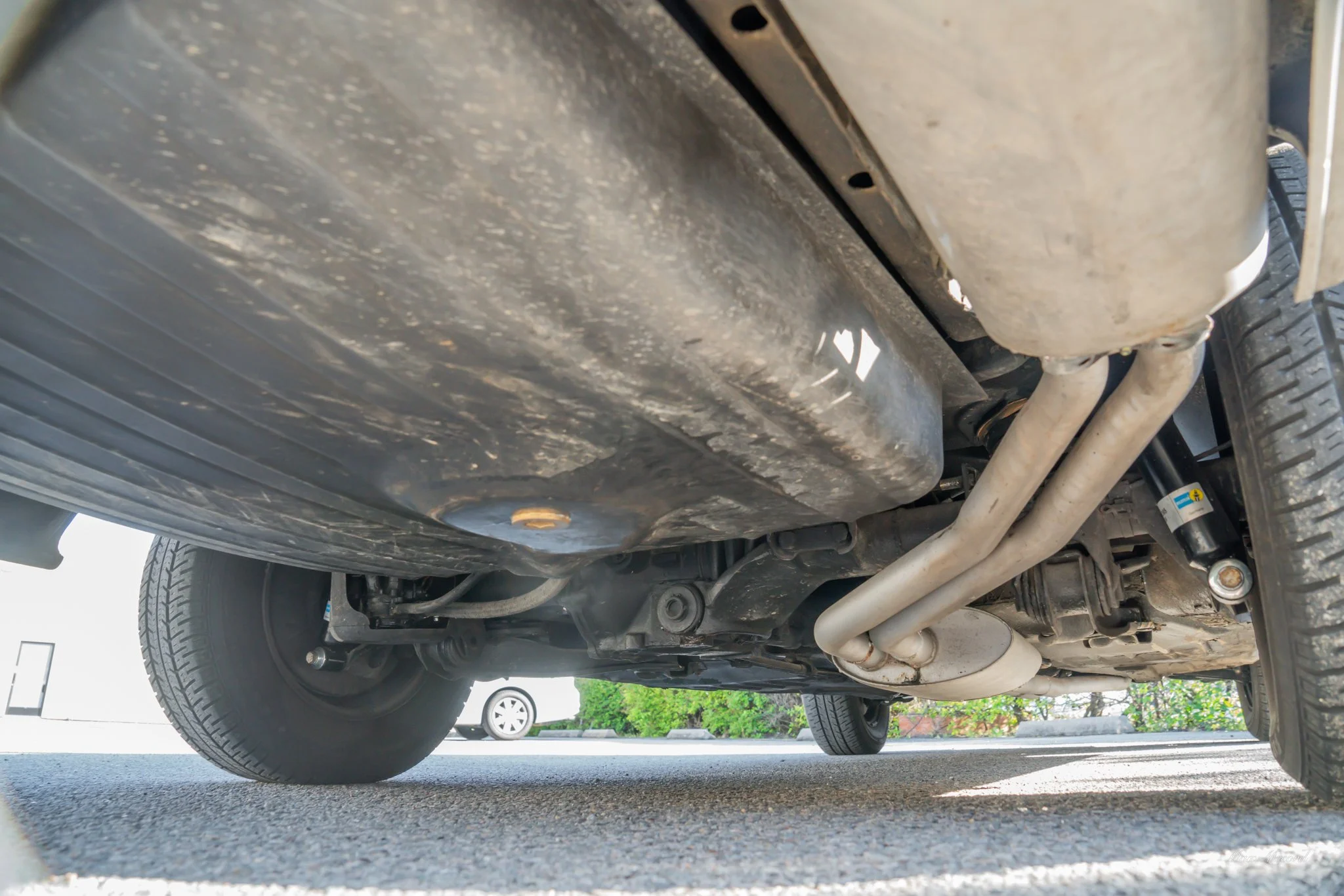Underneath view of a vehicle's exhaust system and oil pan, showing the exhaust pipes and tire on a paved surface.