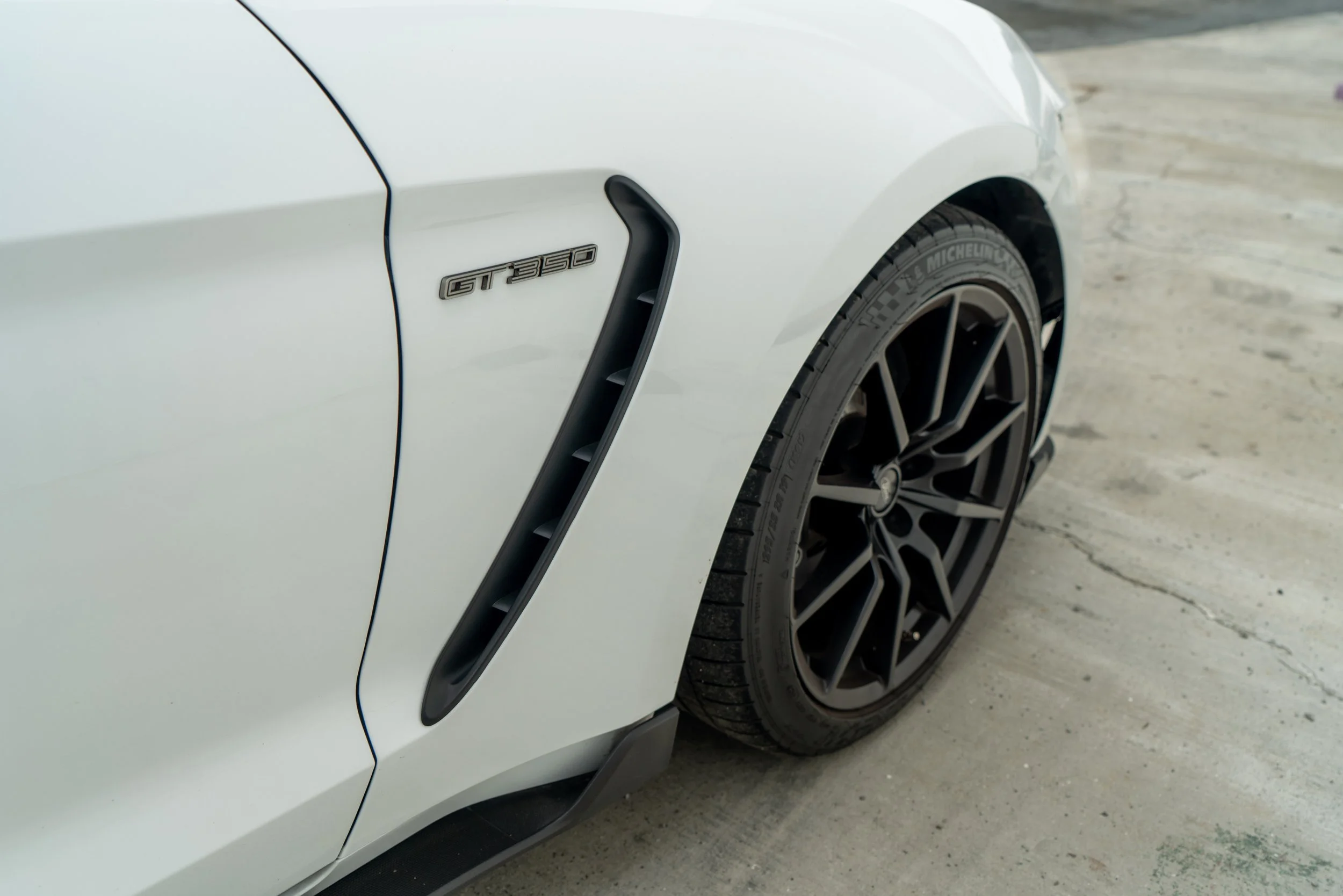 Close-up of a white sports car's front side showing the GT 350 badge, black wheel with Michelin tire, and side vent.