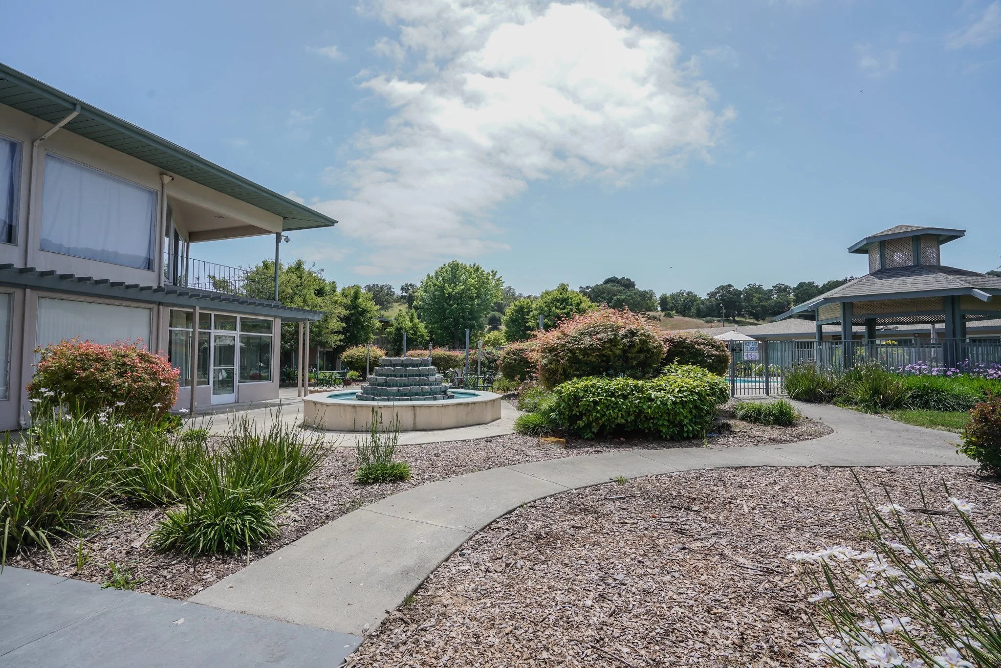 A landscaped outdoor area with a concrete pathway, garden beds with plants and bushes, a water fountain, and buildings with large windows and a gazebo in the background under a partly cloudy sky.