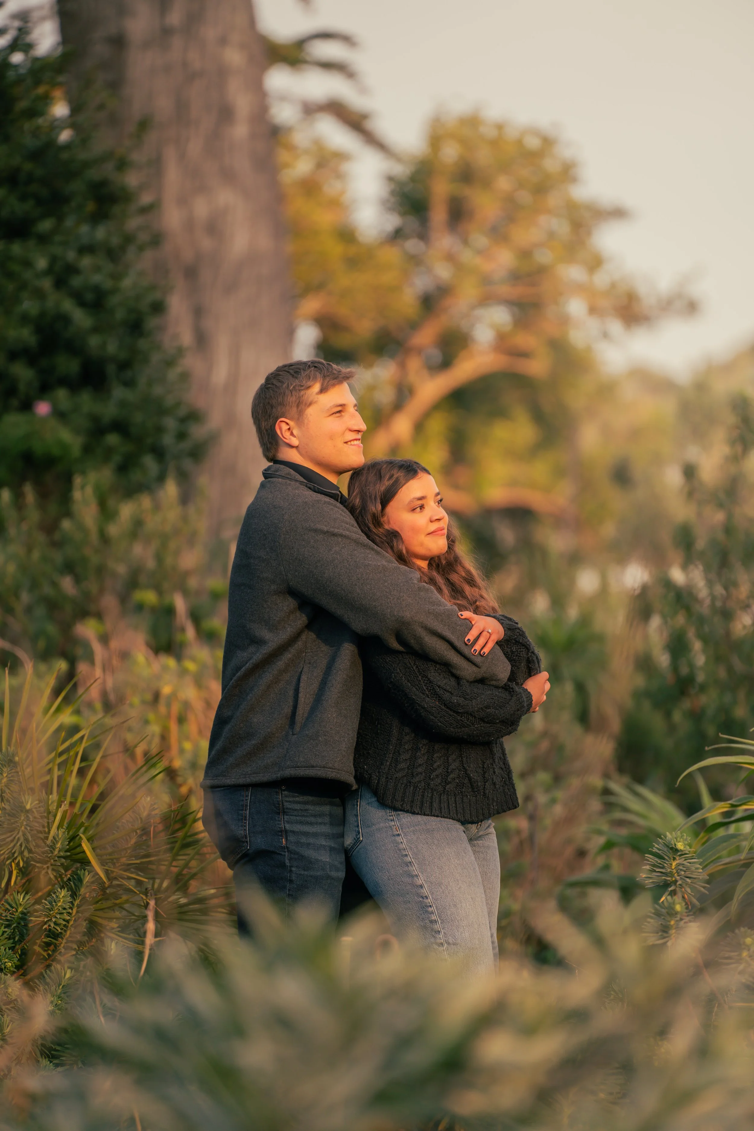 A young couple stands together outdoors, embracing in a natural setting with trees and greenery during sunset.