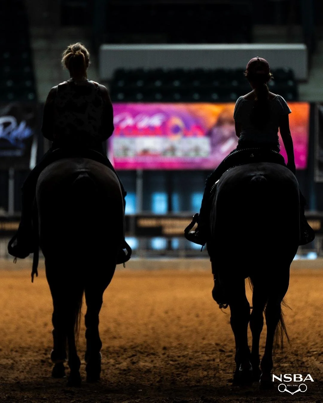 Two women riding horses in an indoor arena at night, silhouetted against a brightly lit digital screen in the background.