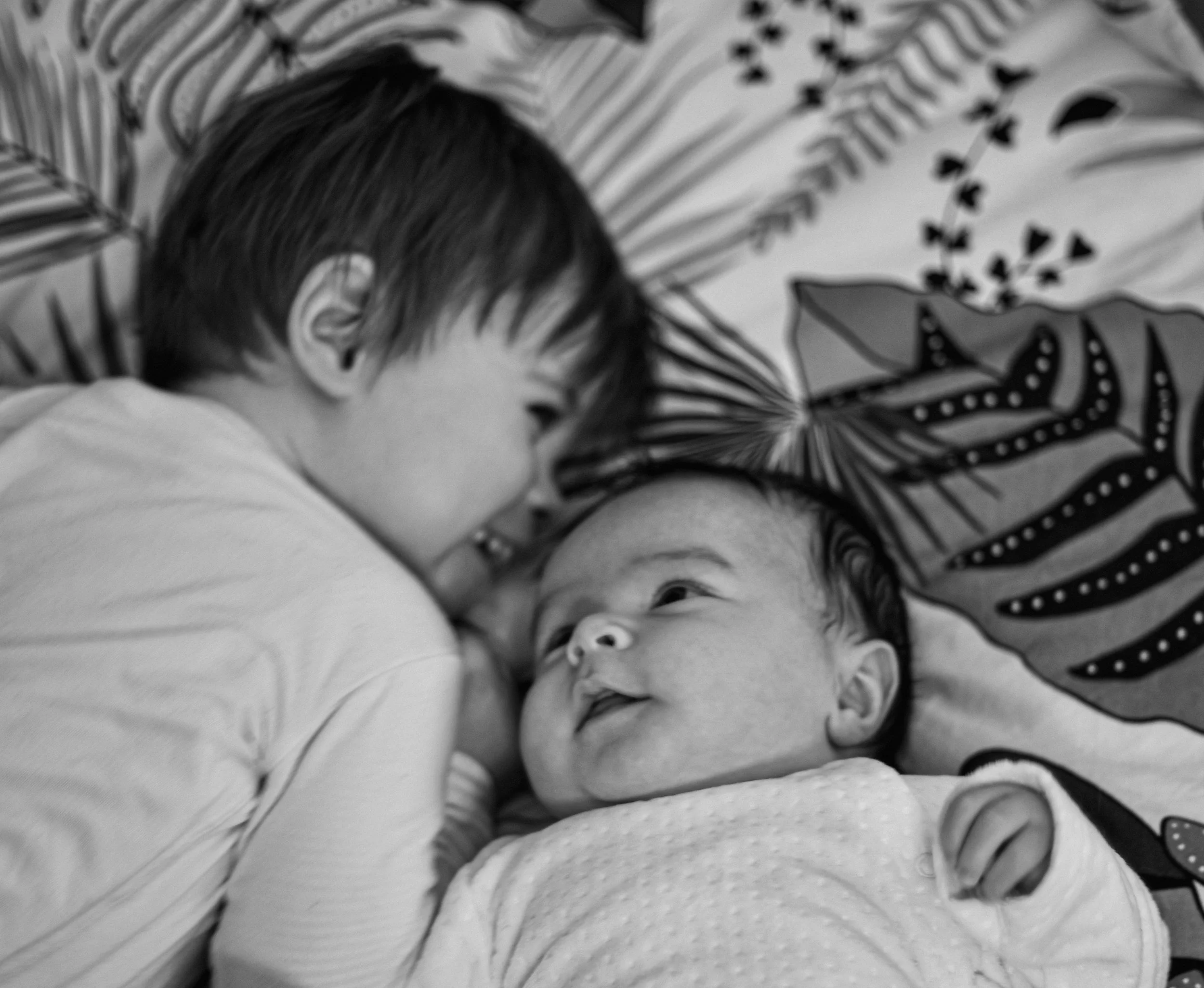 Deux enfants, un plus âgé et un bébé, sourient et regardent l'un l'autre, allongés sur un lit avec des feuilles de monstera imprimées sur les draps.