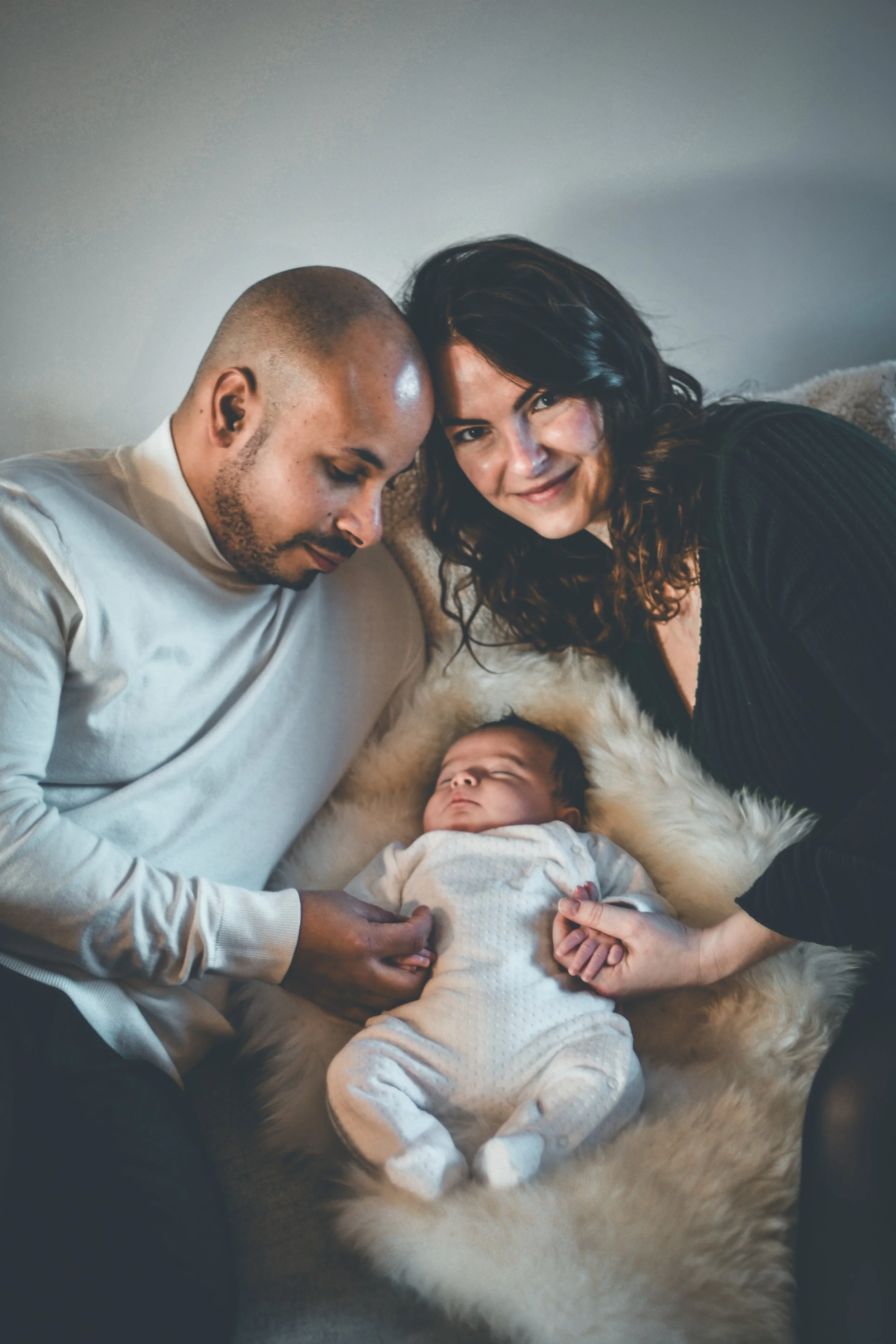Une famille composee d'un homme, d'une femme et de leur bebe endormi sur un couverture en fourrure blanche.