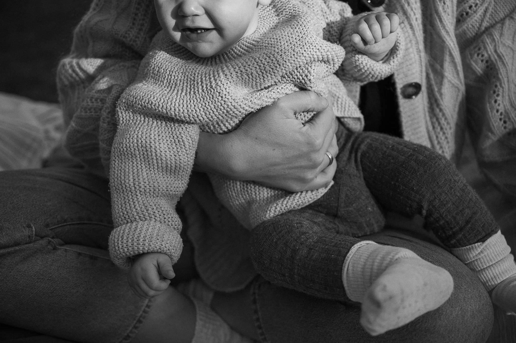 Ein Baby wird von seiner Mutter hochgehoben und lächelt. Das Baby trägt gestrickte Kleidung und Socken, die Mutter hat eine Hand unter dem Baby, während sie vermutlich lachen oder sprechen.