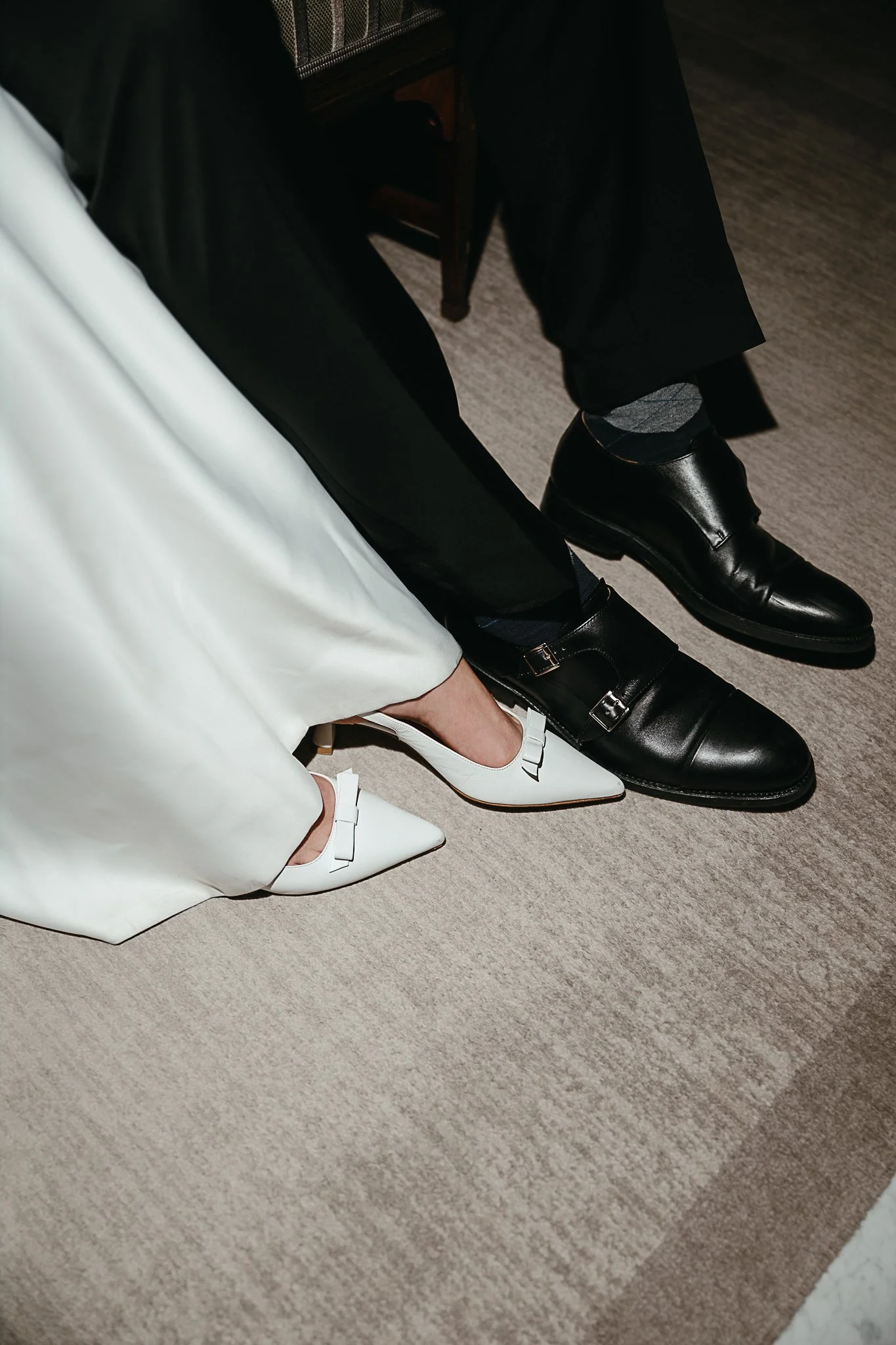 Nahaufnahme eines Brautpaares in Weiß mit weißen Slingback-Pumps und einem schwarzen Lederschuhen auf einem Teppich.