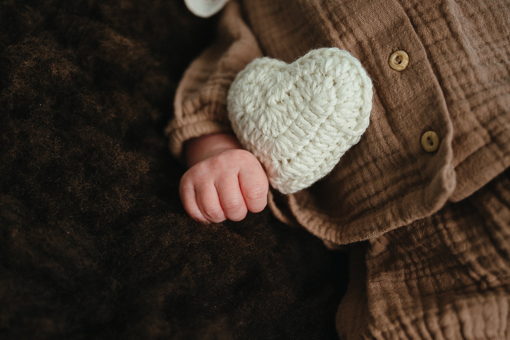 Nahaufnahme eines Babys in brauner Kleidung mit gestricktem weißen Herz und kleiner Hand auf einer dunklen Decke.