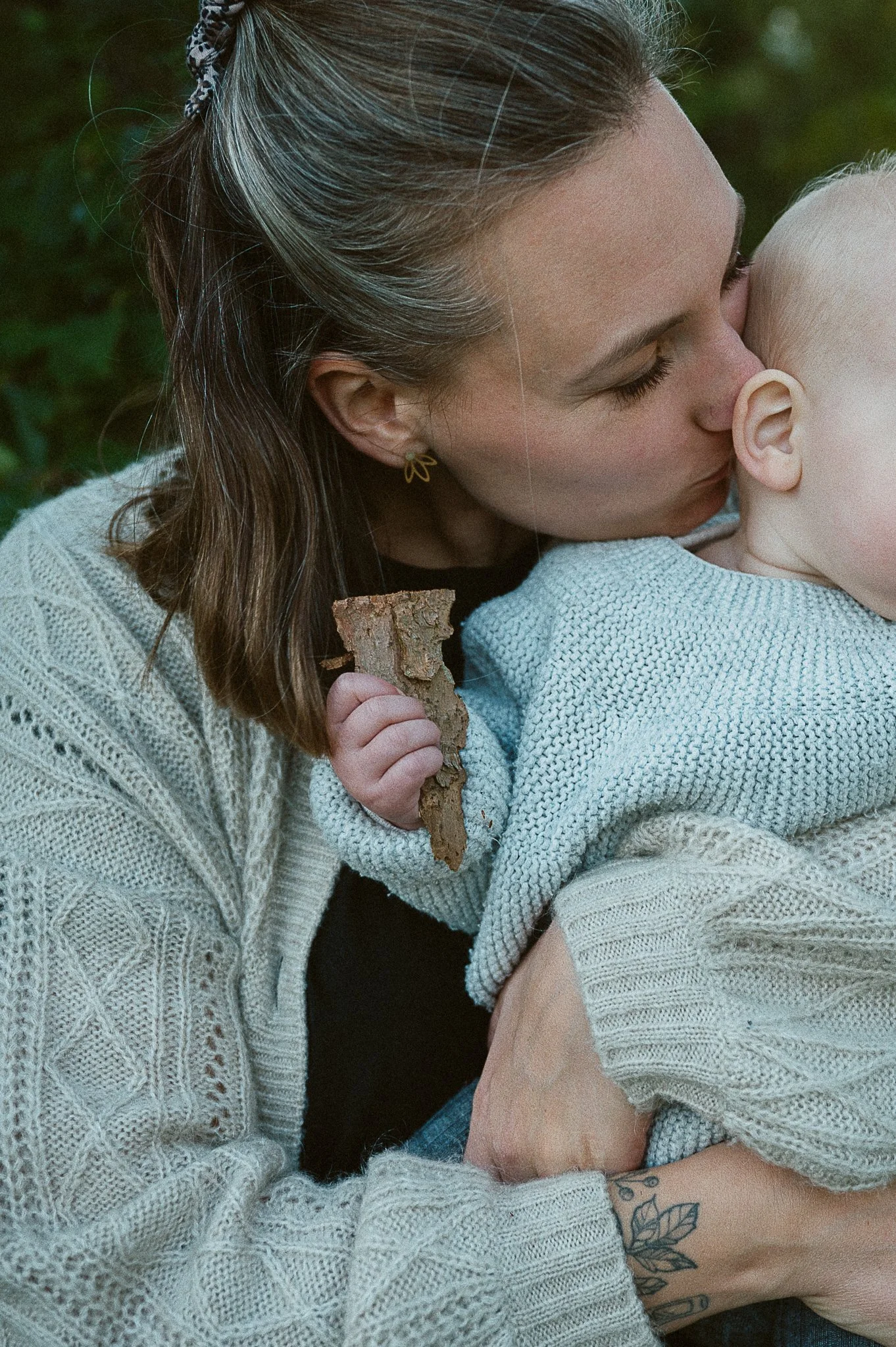 Eine Frau hält ein Baby im Arm und küsst es auf den Kopf, sie sitzen in einem Garten.