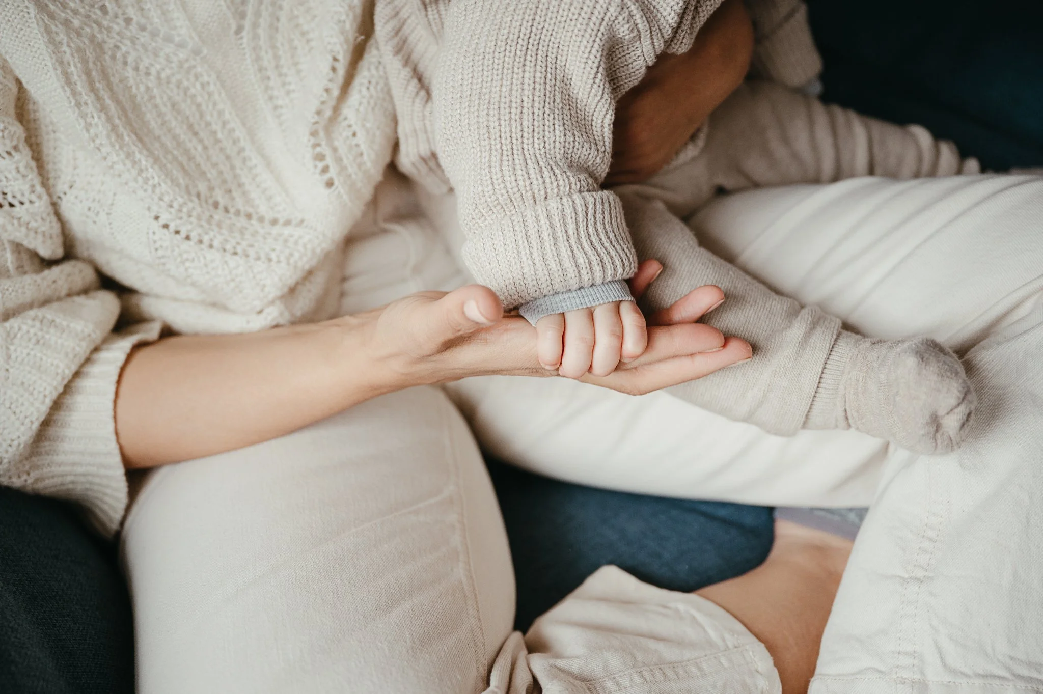 Eine erwachsene Person hält die Hand eines Kindes, das in einem zarten Grau gekleidet ist, während sie zusammen sitzen.
