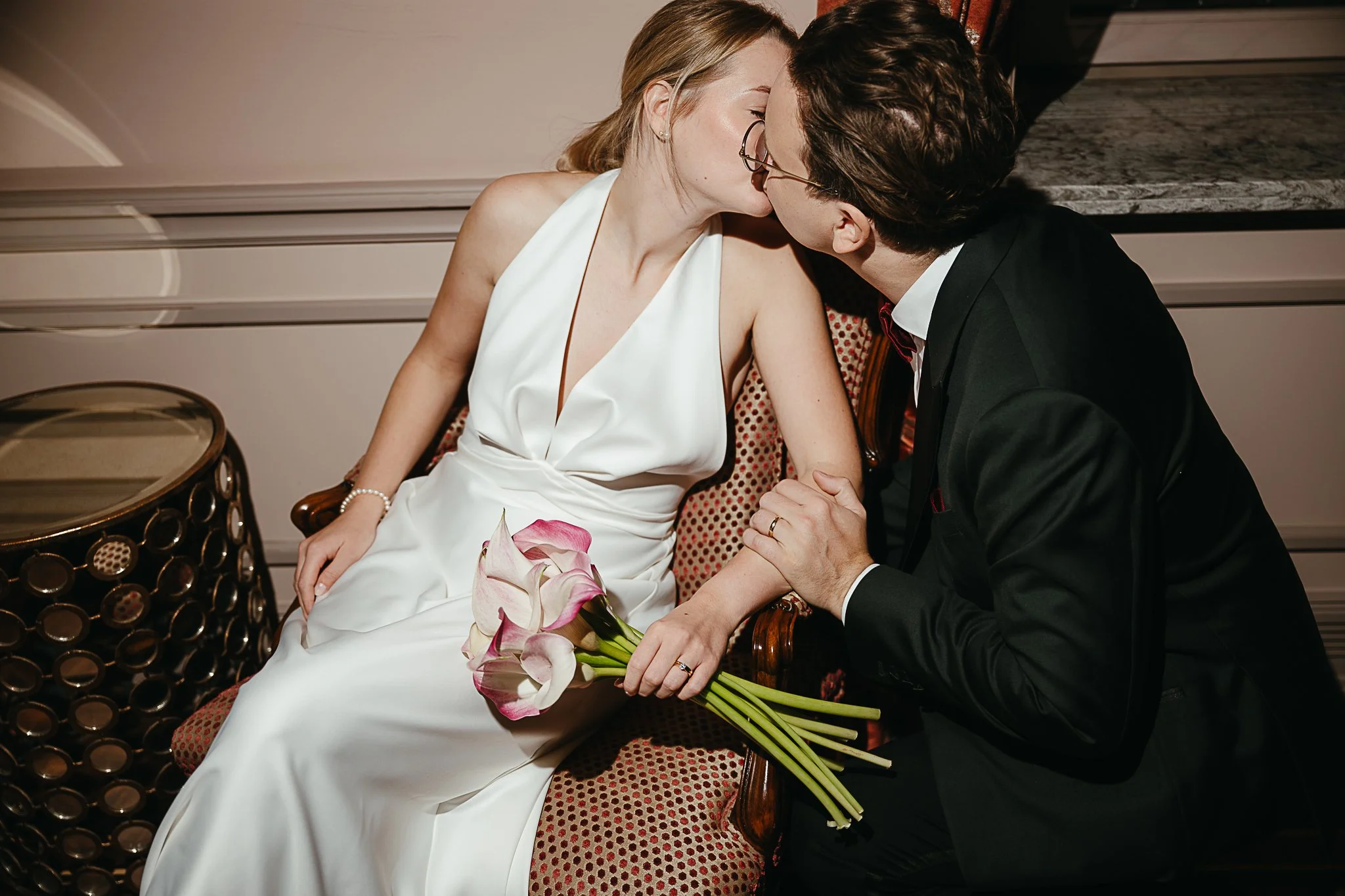 Brautpaar küsst sich in einem Sessel; die Frau im Hochzeitskleid hält einen Brautstrauß mit Calla-Lilien, der Mann trägt einen schwarzen Hochzeitsanzug.