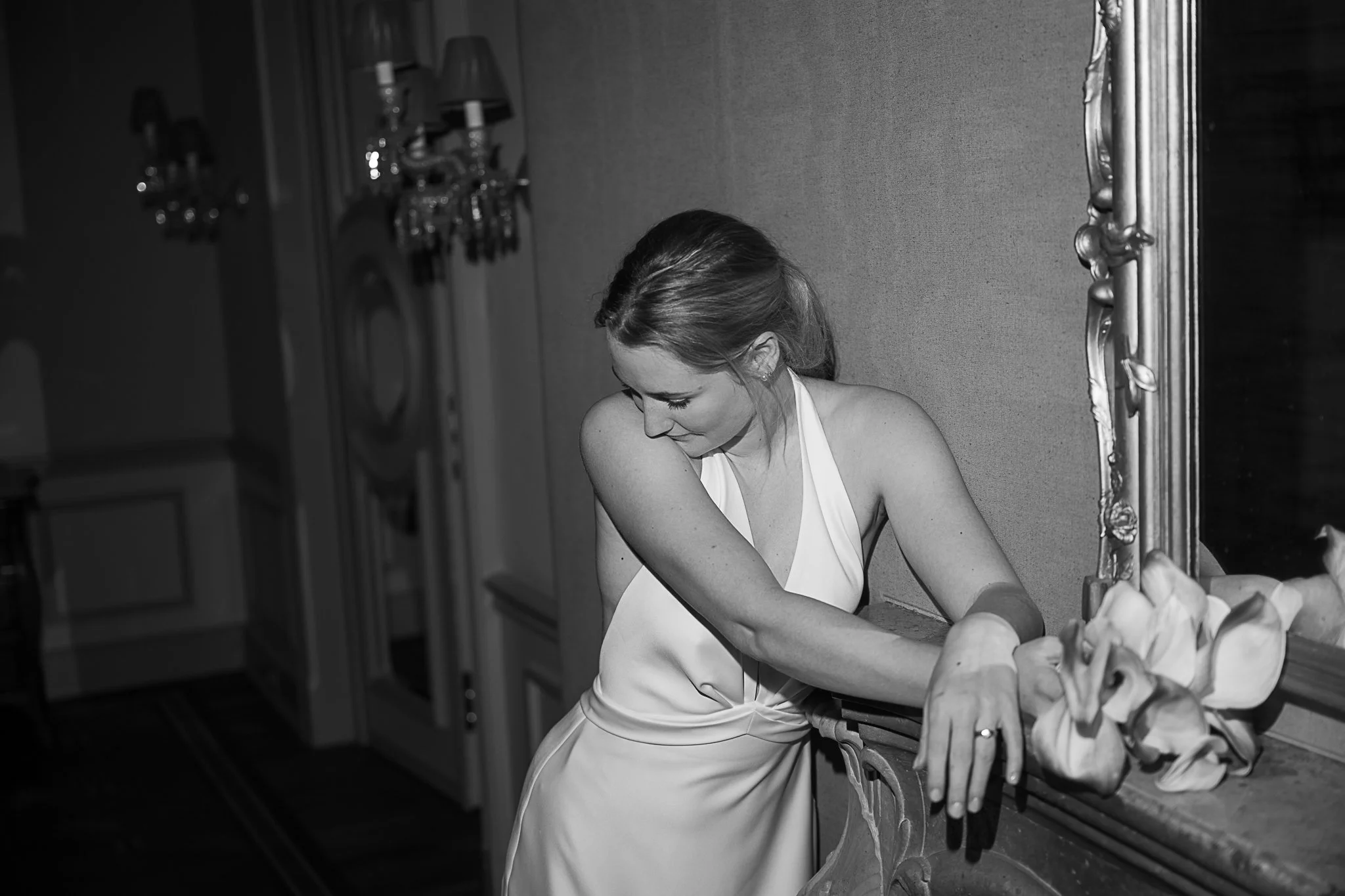 Schwarz-Weiß-Foto einer Braut im Hochzeitskleid, die sich an einen Kaminsims mit Spiegel lehnt.