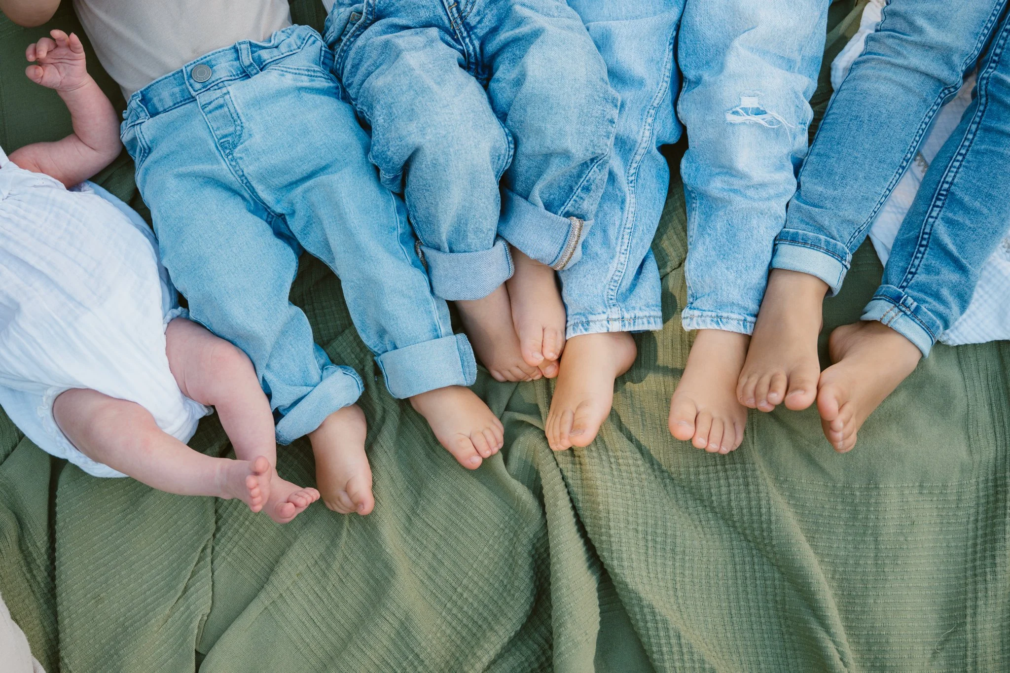 Fünf Kinder und ein Baby mit unterschiedlichen Jeanshosen, die nebeneinander auf einer grünen Decke liegen.