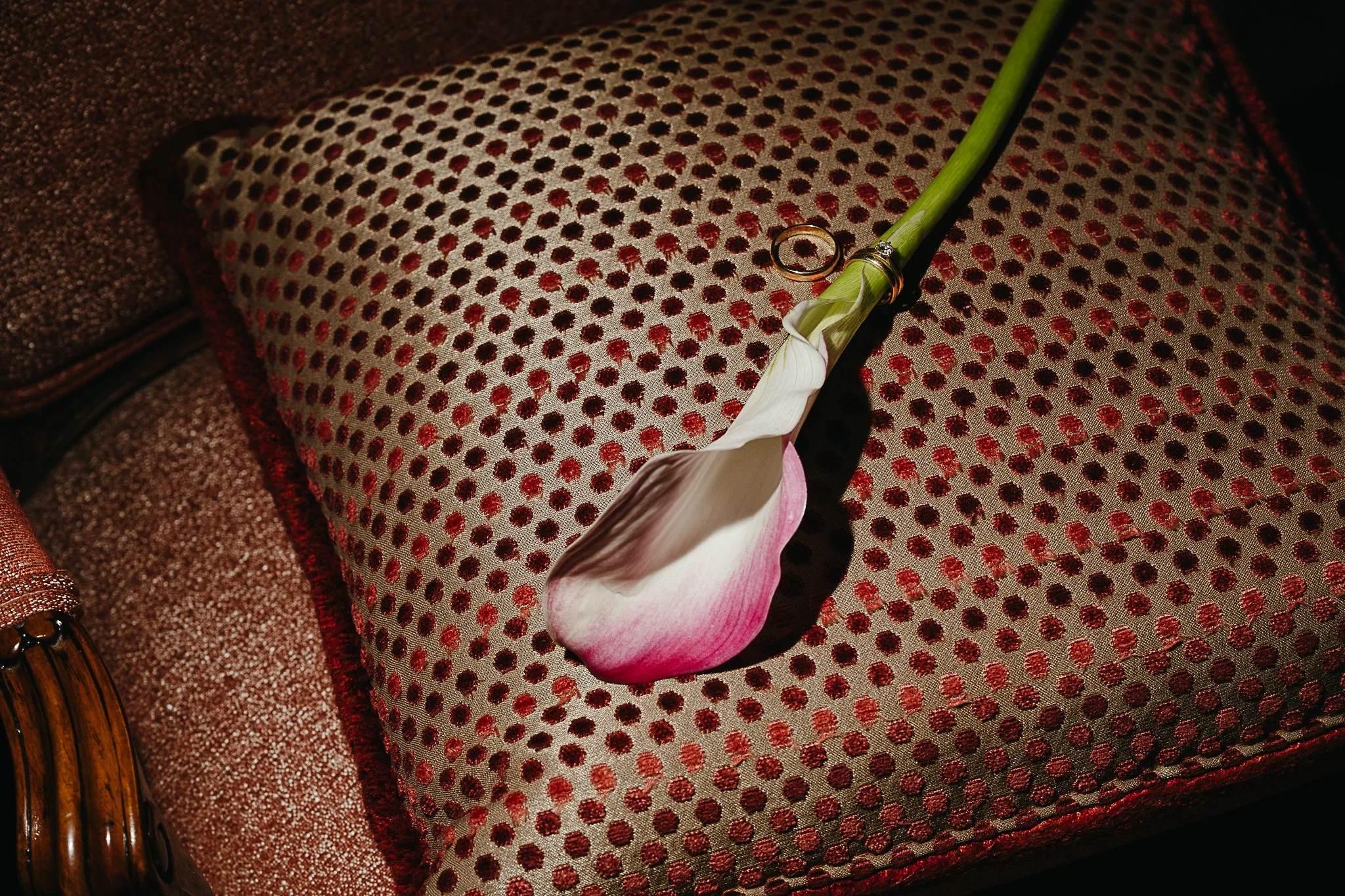 Detailbild einer weiß-rosa Calla-Lilie aus einem Brautstrauß mit goldenen Eheringen auf einem rot gemusterten Polstersessel