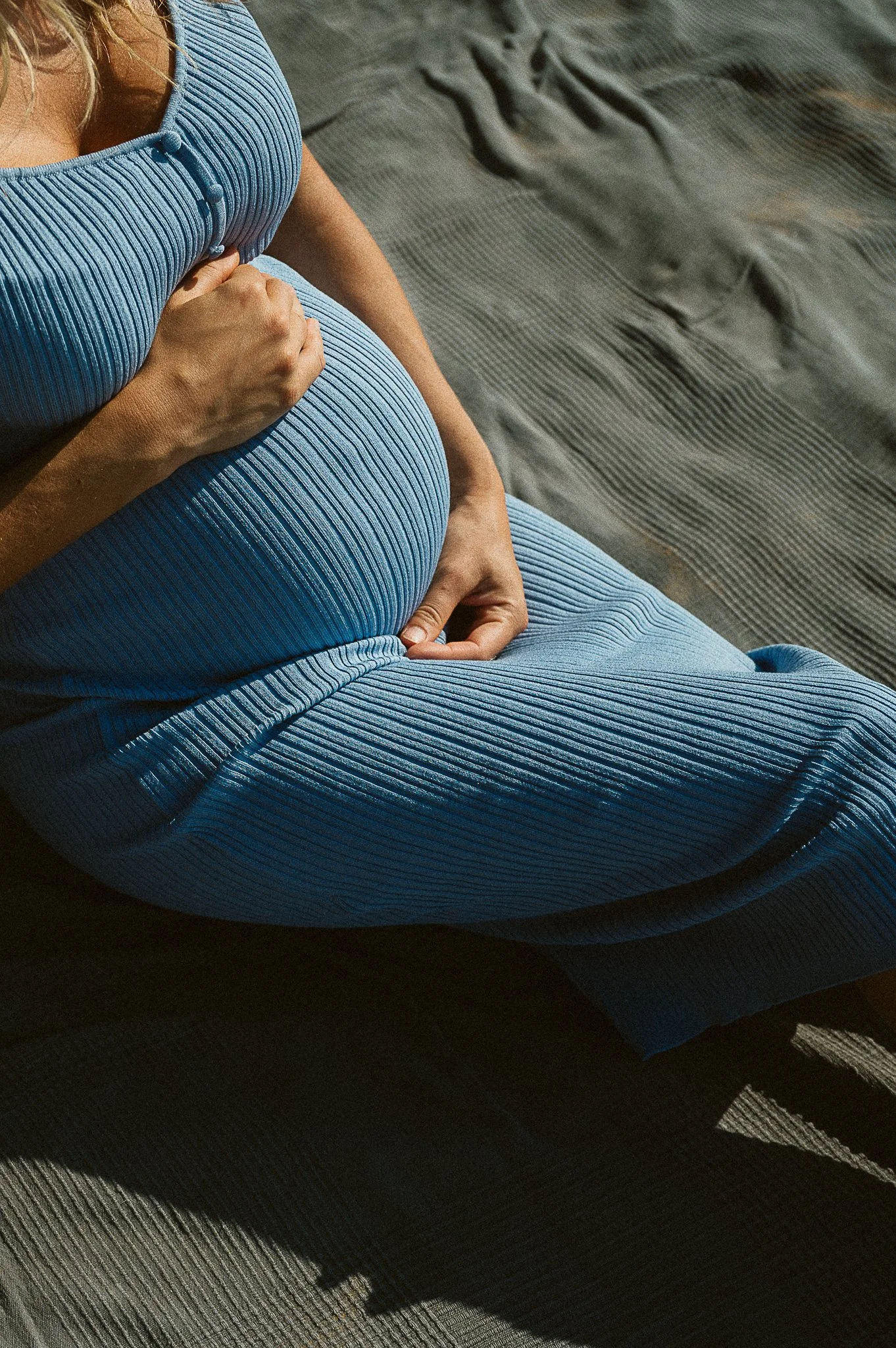 Nahaufnahme einer schwangeren Frau in einem blauen, gerippten Kleid, die auf einer grauen Decke liegt. Ihre Hand ruht auf ihrem Babybauch.