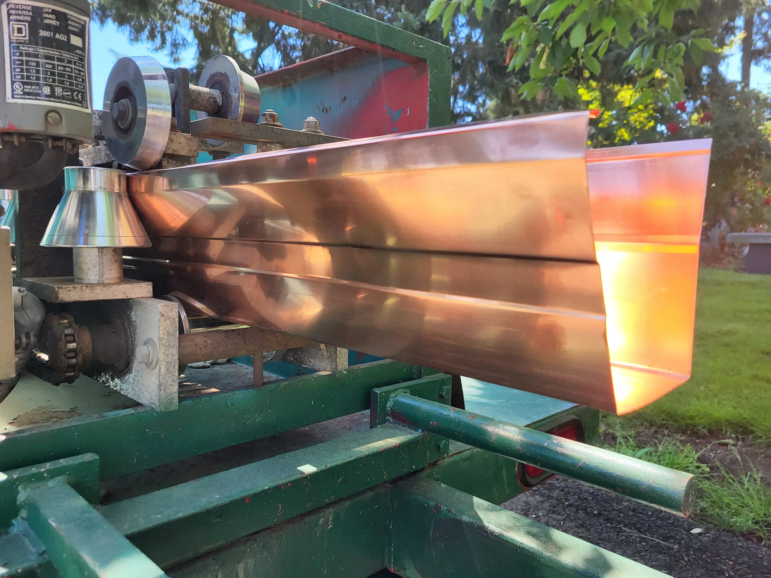 A wide bottom gutter machine, mounted on a green frame, is outdoors with trees and blue sky in the background.