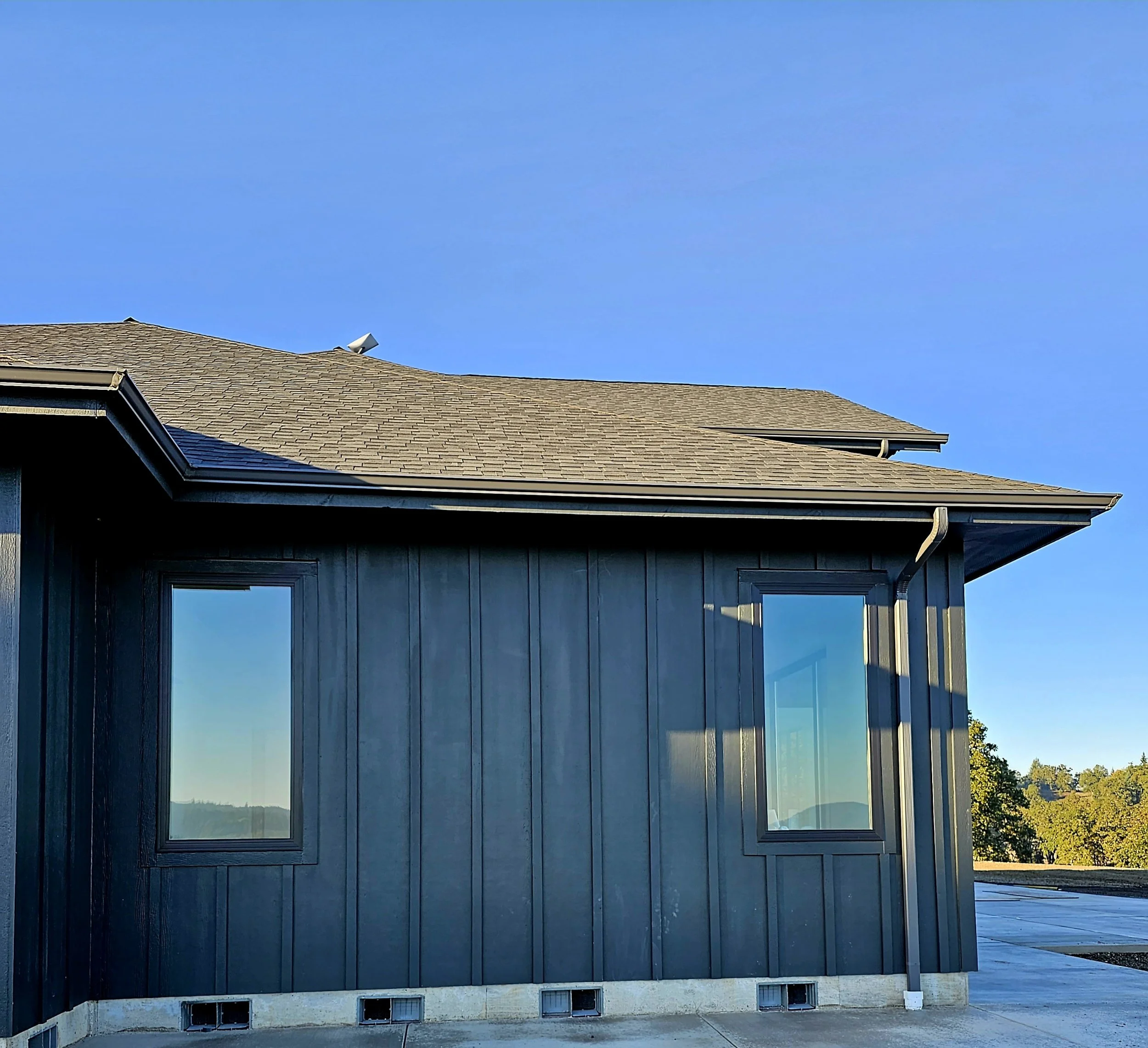 View of a modern house with large glass windows, gray siding, and a sloped shingled roof against a clear blue sky.