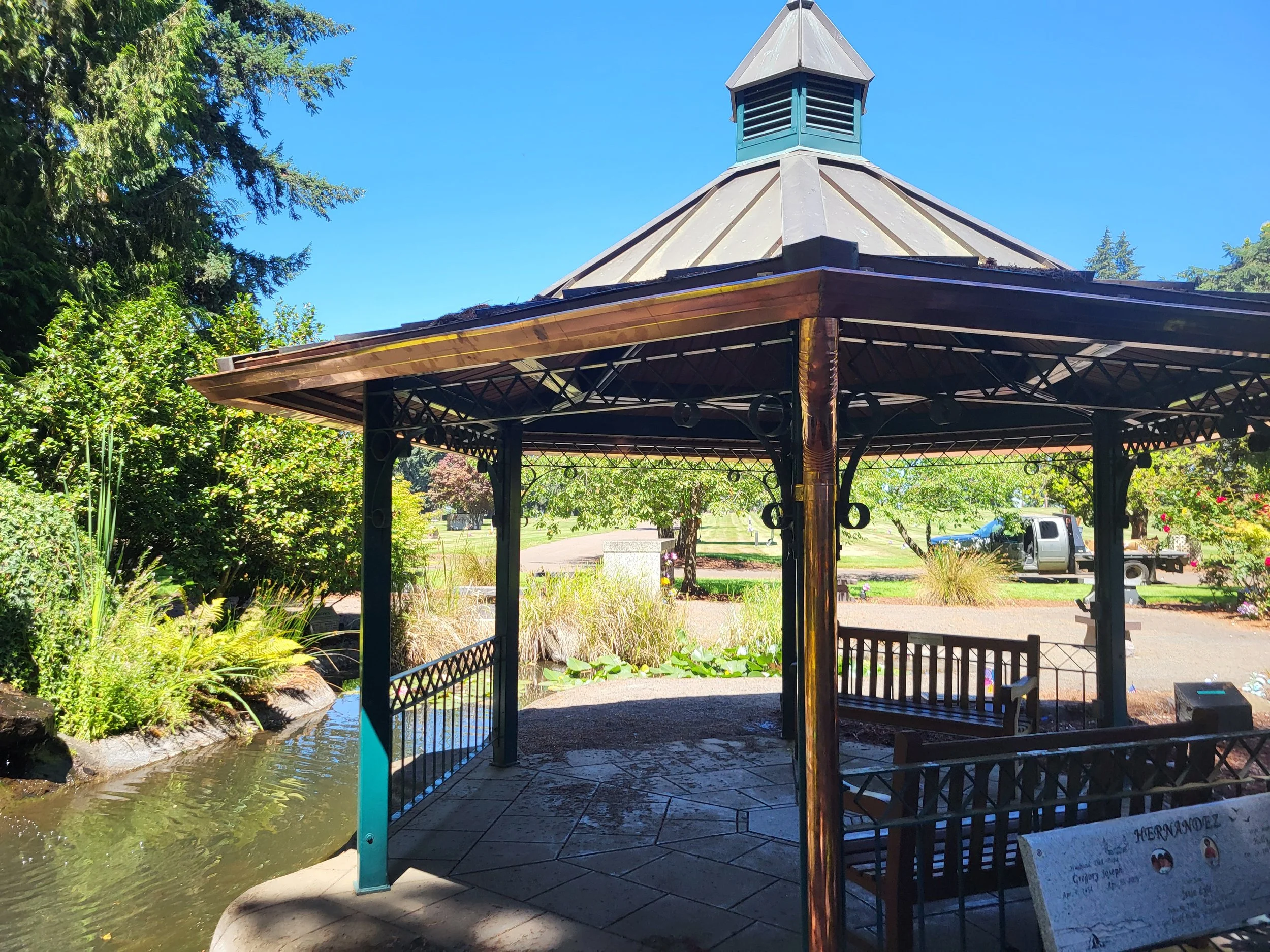 A gazebo with a copper roof  and gutters in a park, surrounded by greenery and trees, with a small pond nearby and a clear blue sky above.