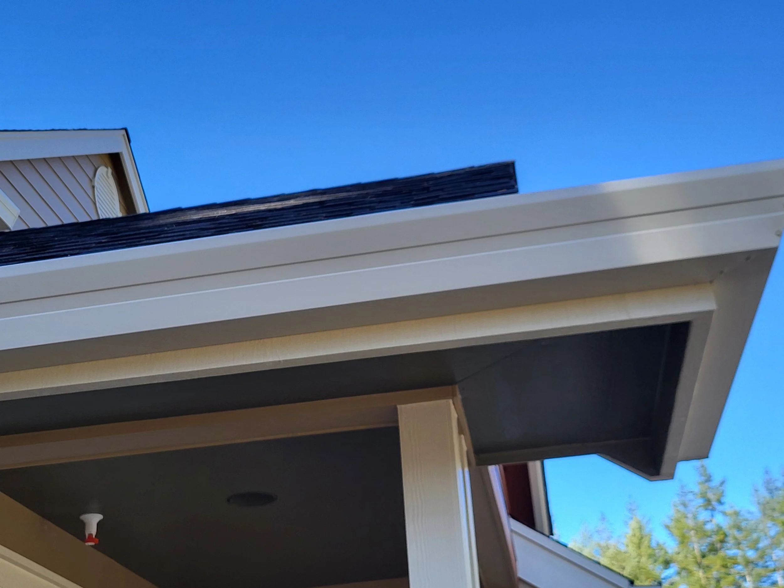 A view of a house exterior showing a white porch ceiling, part of a wall with beige siding, and an open window with a white frame. The sky is clear and blue with some green trees visible in the background.