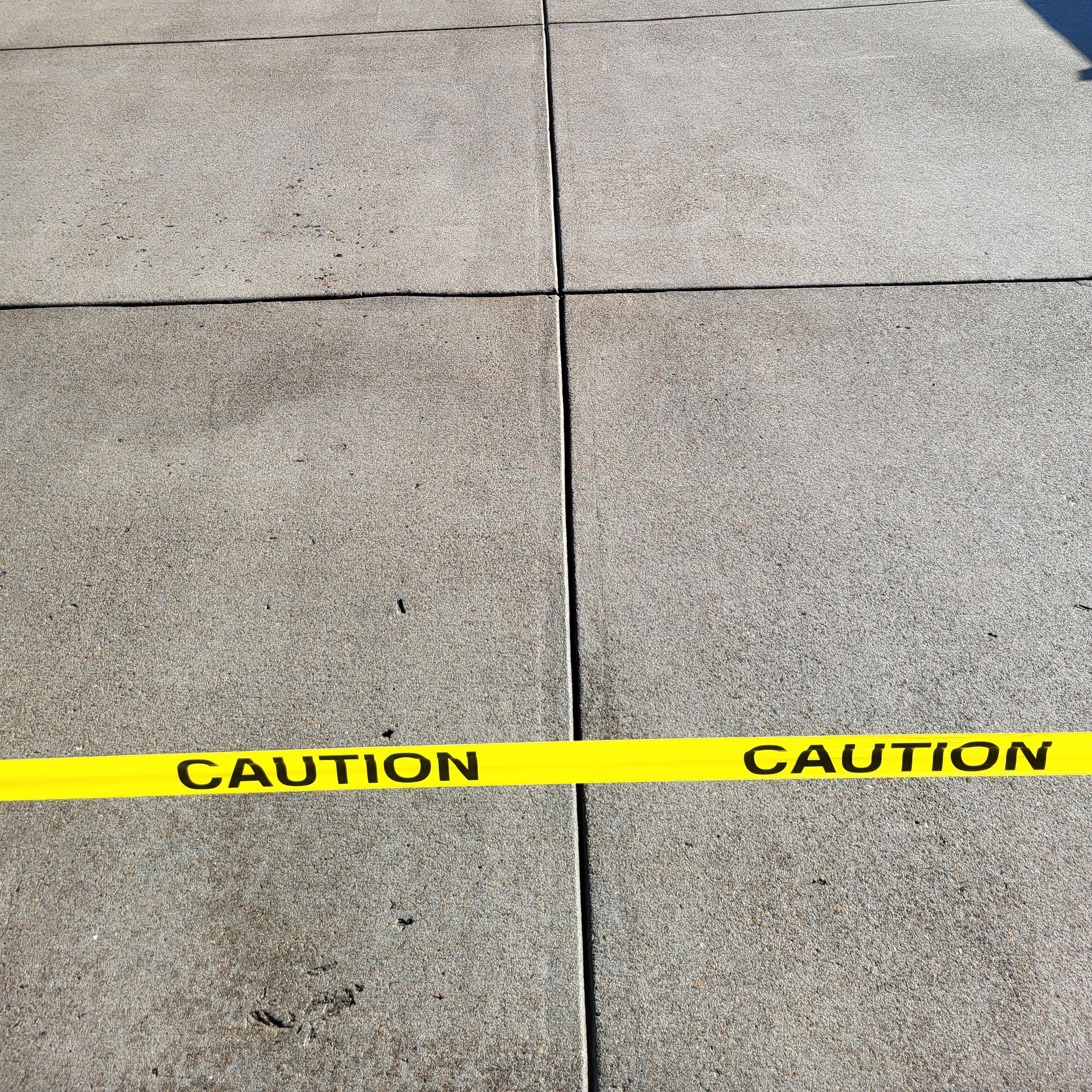 Concrete sidewalk with four tiles, a corner of yellow caution tape at the bottom, and a shadow cast on the top right corner.