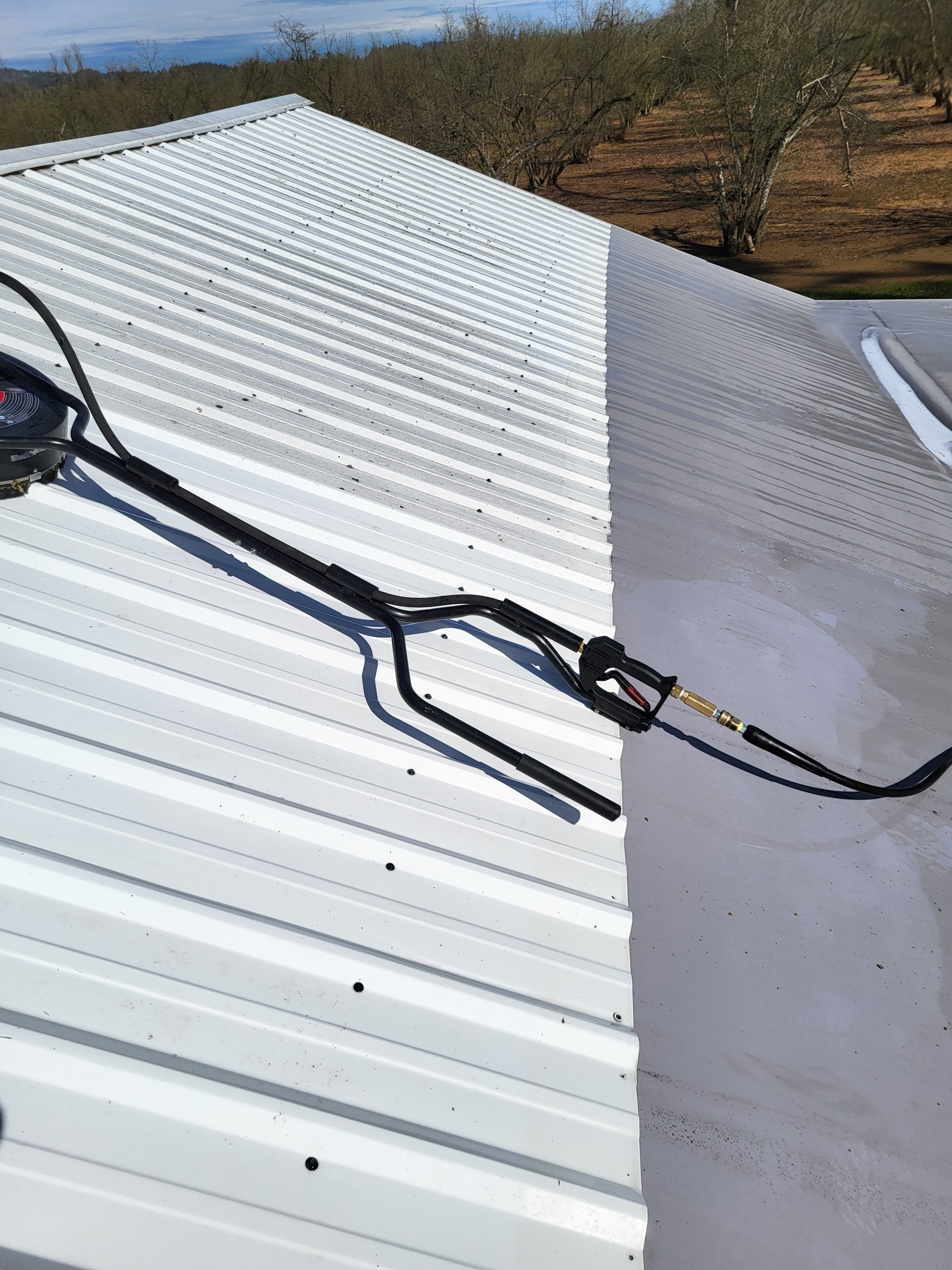 Metal roof with a power washing brush, set against a background of trees and a clear sky.