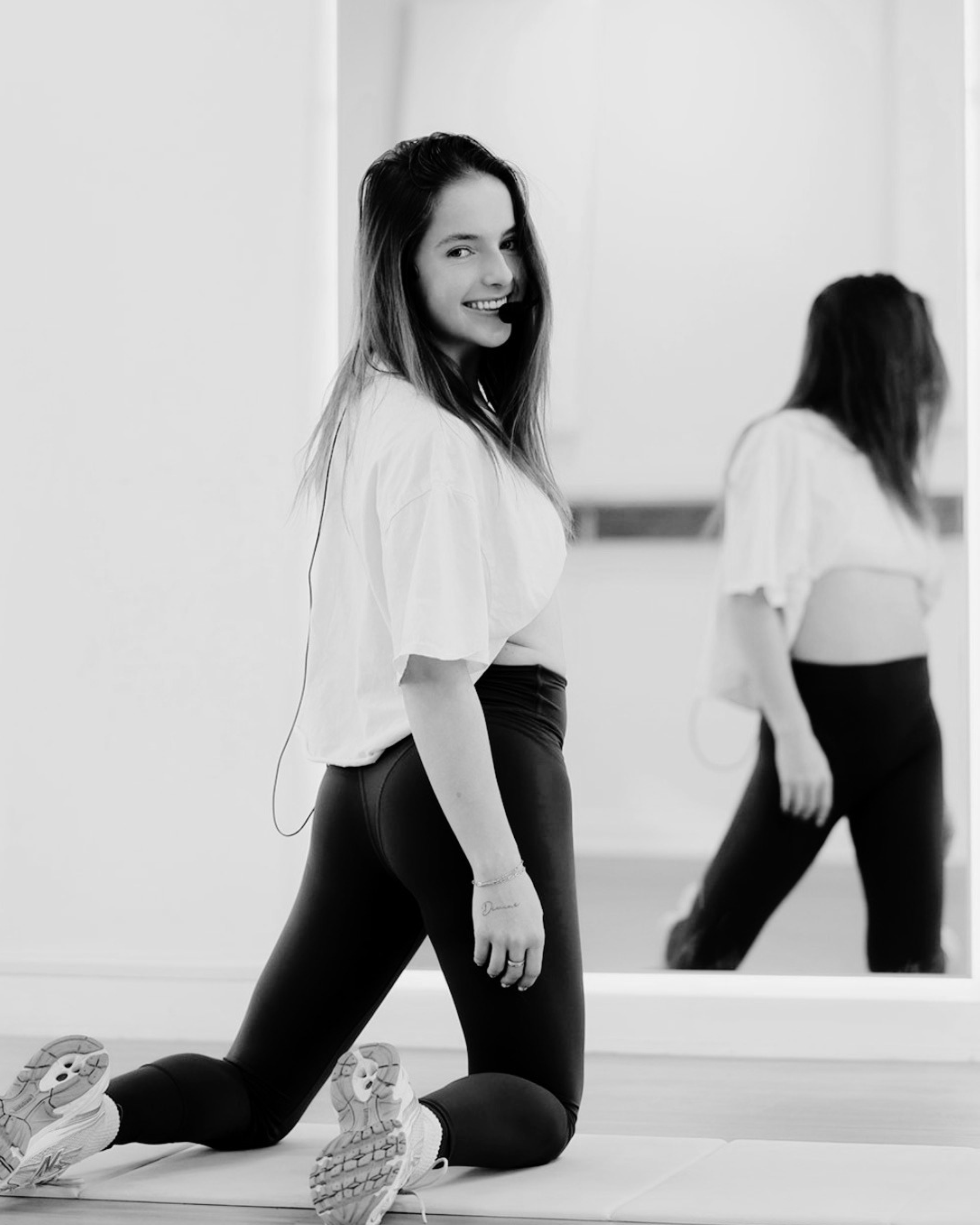 A young woman in athletic clothing, wearing a microphone headset, is kneeling on a mat in a fitness studio. She is smiling and looking at the camera with her right hand hanging down. Her reflection appears in a large mirror behind her, and another woman in workout clothes is seen in the mirror, also participating in the fitness session.
