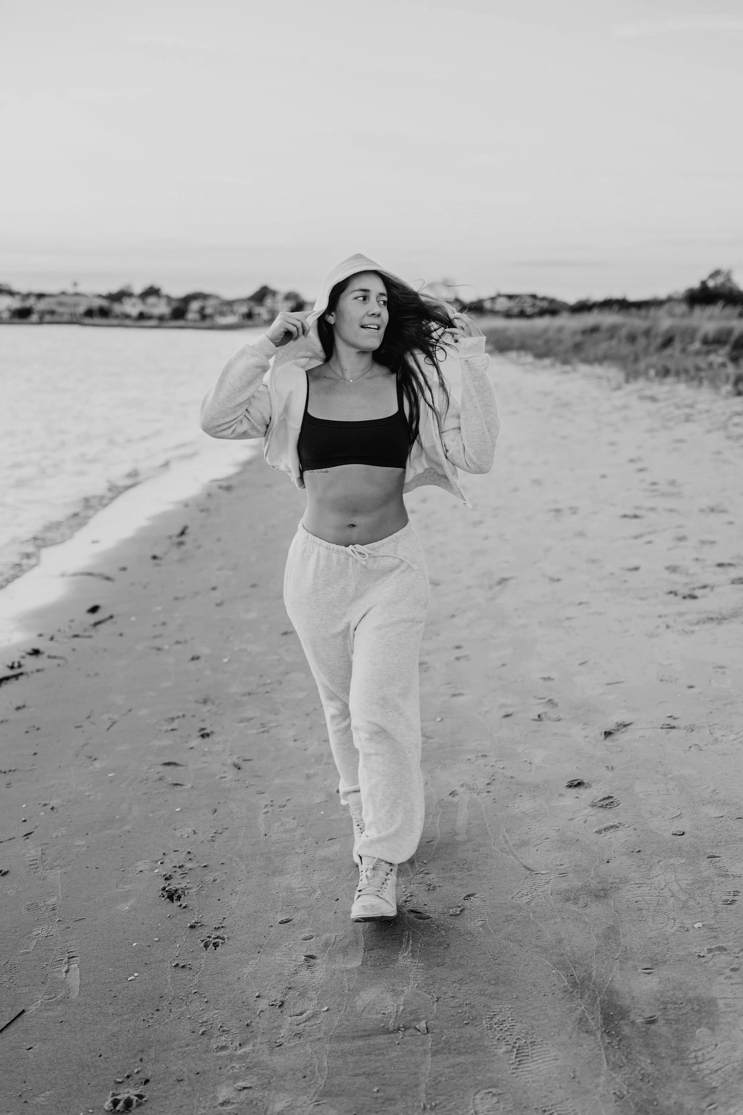 A woman jogging on a sandy beach, wearing a hoodie, sweatpants, and a sports bra, with her hair flowing in the wind.