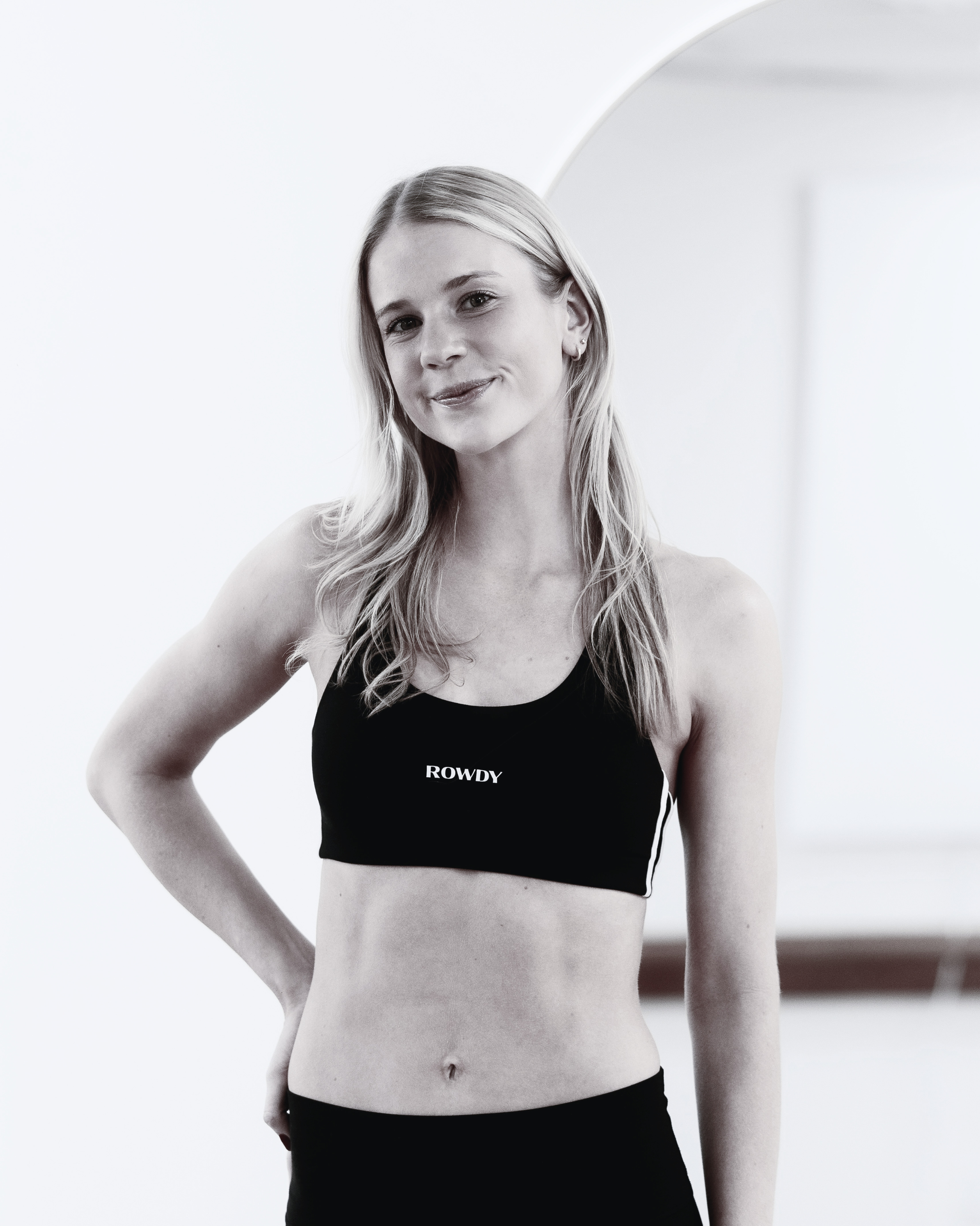 A young woman with long wavy blonde hair wearing a black sports bra with the word 'ROWDY' on it, standing with one arm on her hip, smiling slightly, in a minimalist room with white walls.