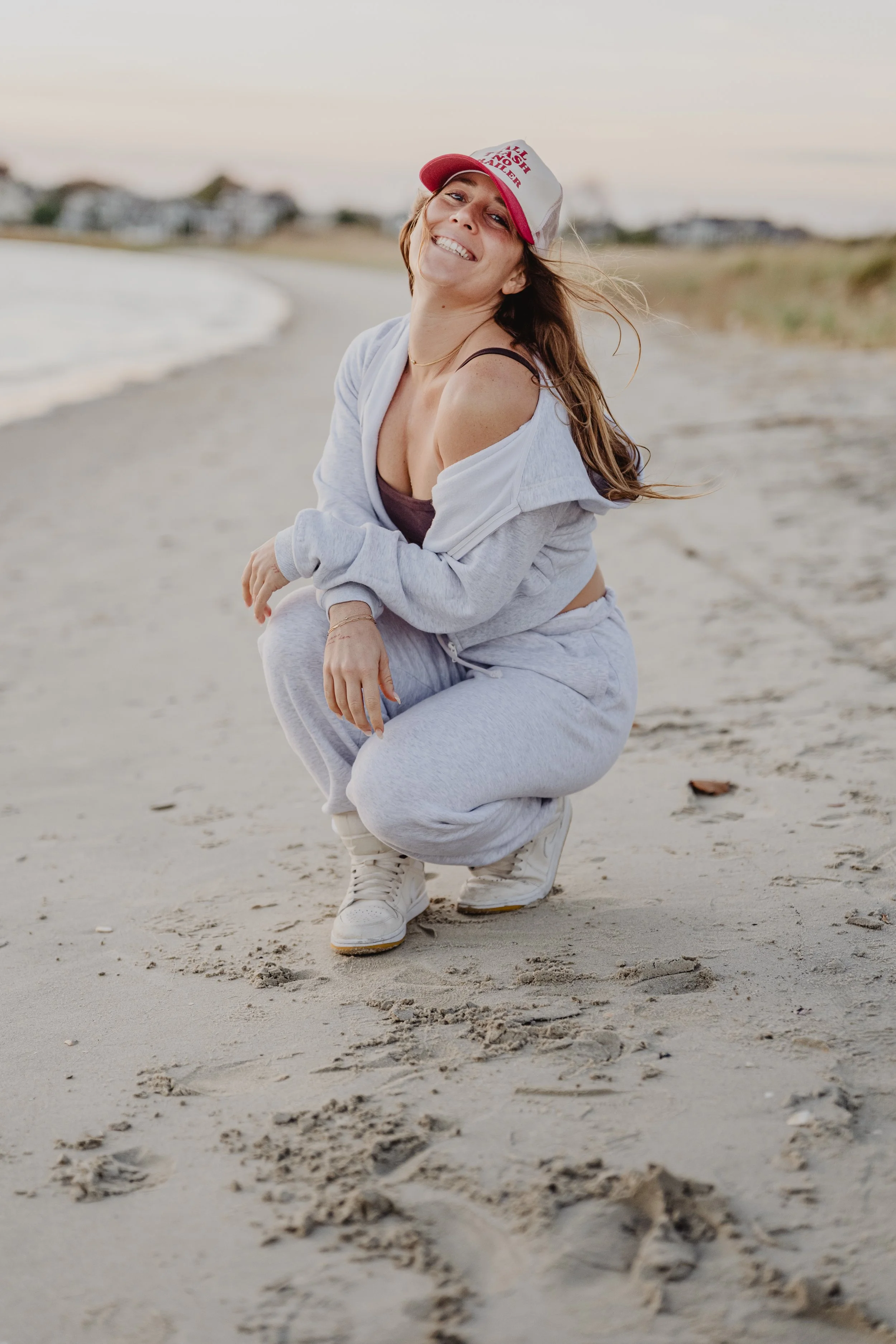 A woman smiling and squatting on a sandy beach, wearing a gray hoodie, gray sweatpants, white sneakers, and a white and red trucker hat, with wind blowing her hair.