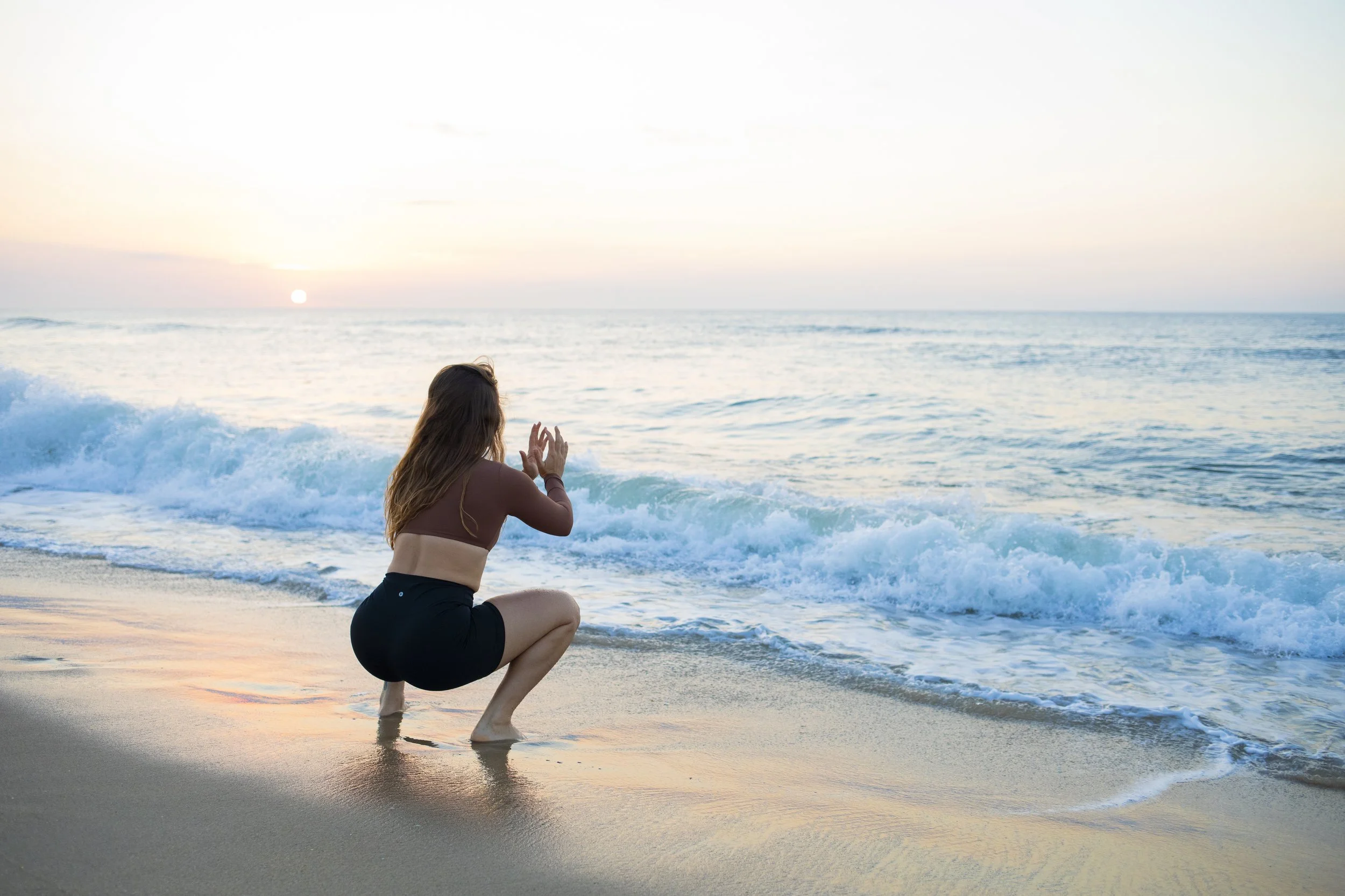 A woman with long hair, wearing a sports bra and shorts, crouching on the beach near the waves with a sunset in the background.