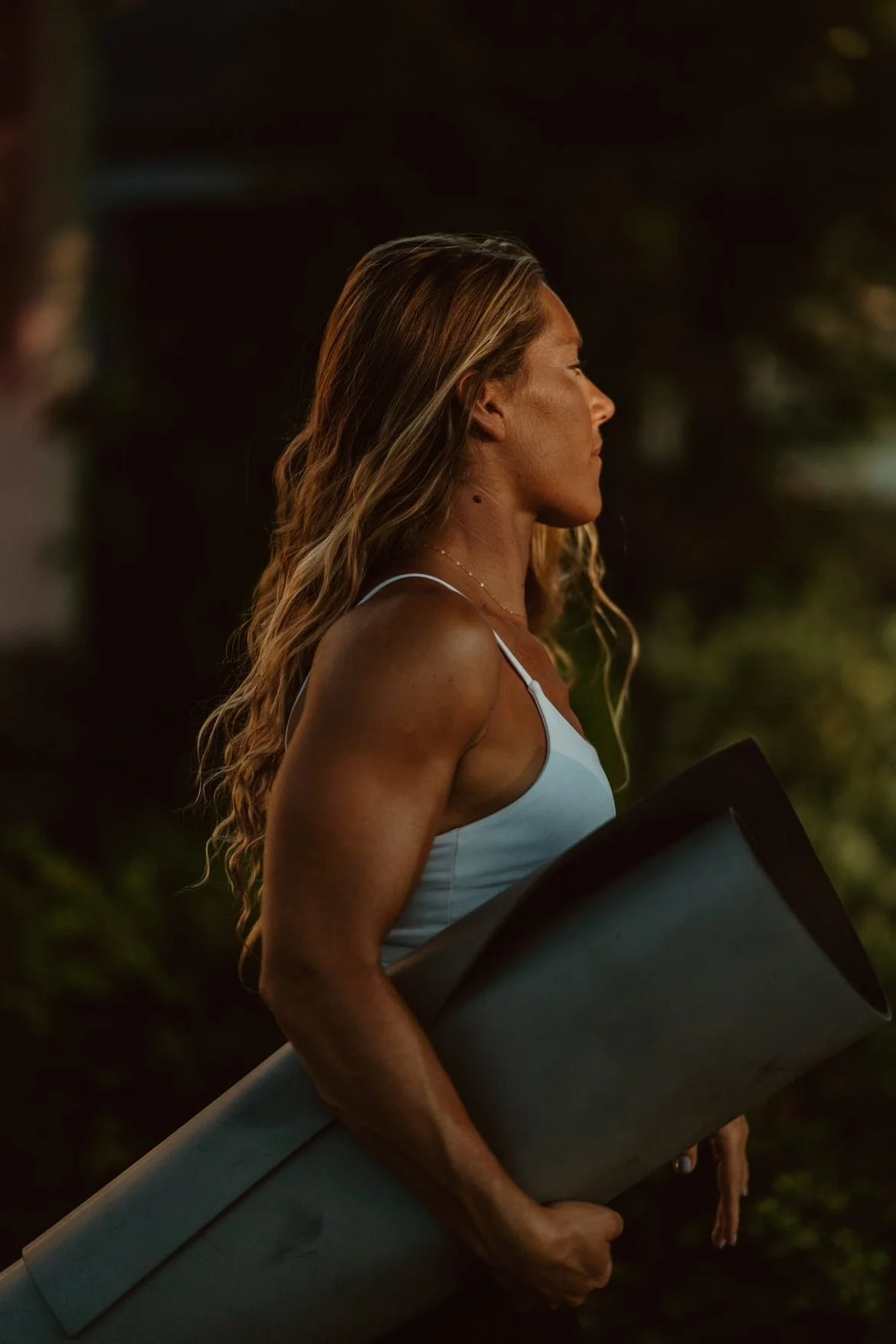 A woman with wavy blonde hair and tanned skin walking outdoors at sunset, holding a rolled-up yoga mat.