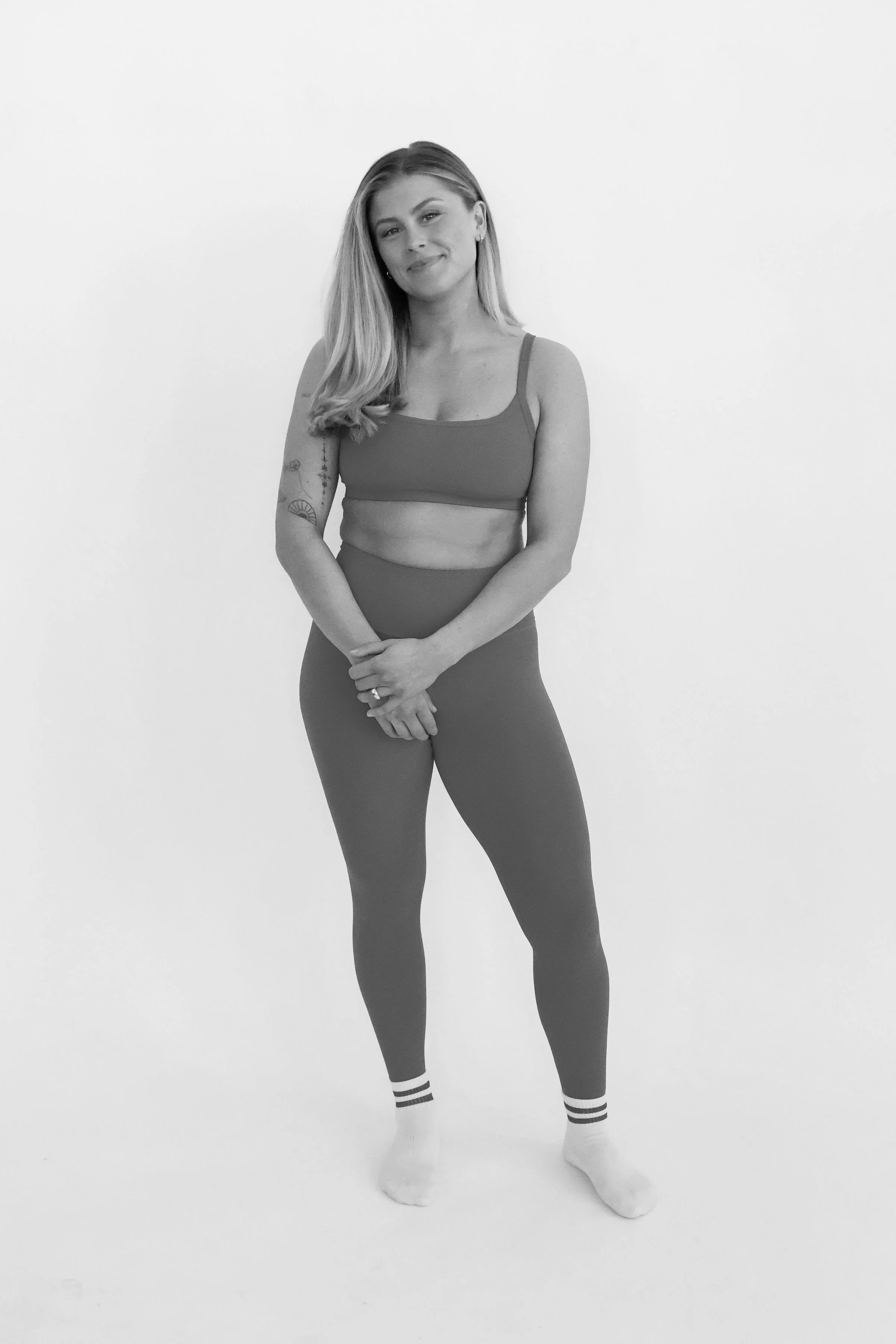 A woman with long hair and tattoos standing against a plain wall, wearing a sports bra, leggings, and socked feet, smiling slightly.