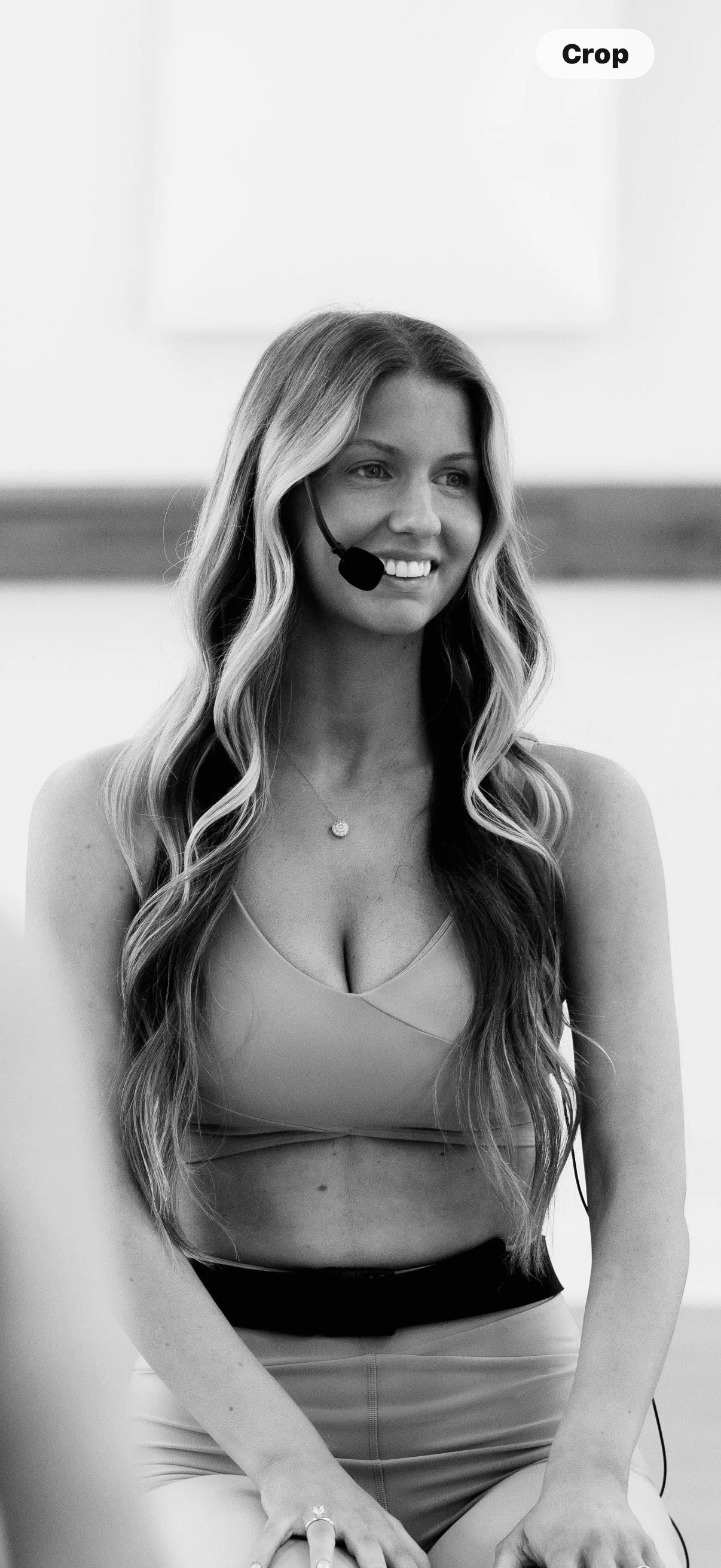 A woman with long wavy hair wearing a headset, a tank top, and a necklace, sitting and smiling in an indoor setting.