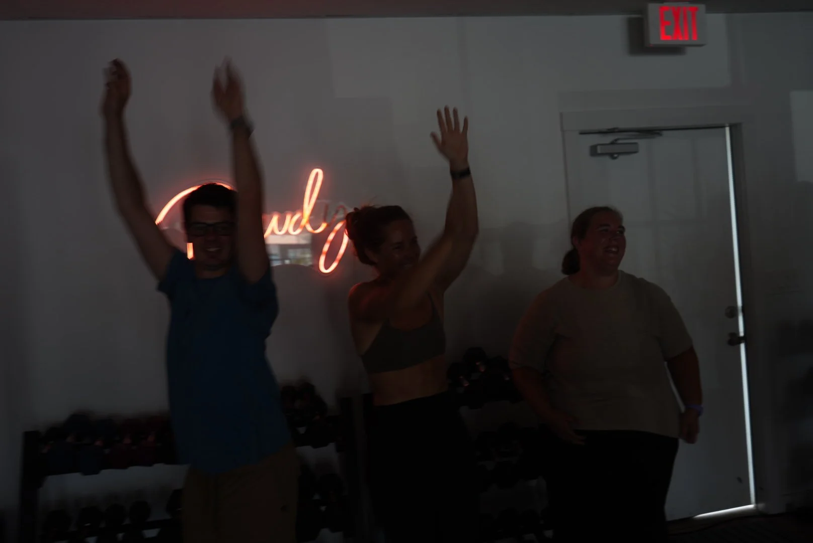 Three women celebrating indoors with raised hands, smiling, in front of a neon sign that says 'Good Vibes' and an exit door.