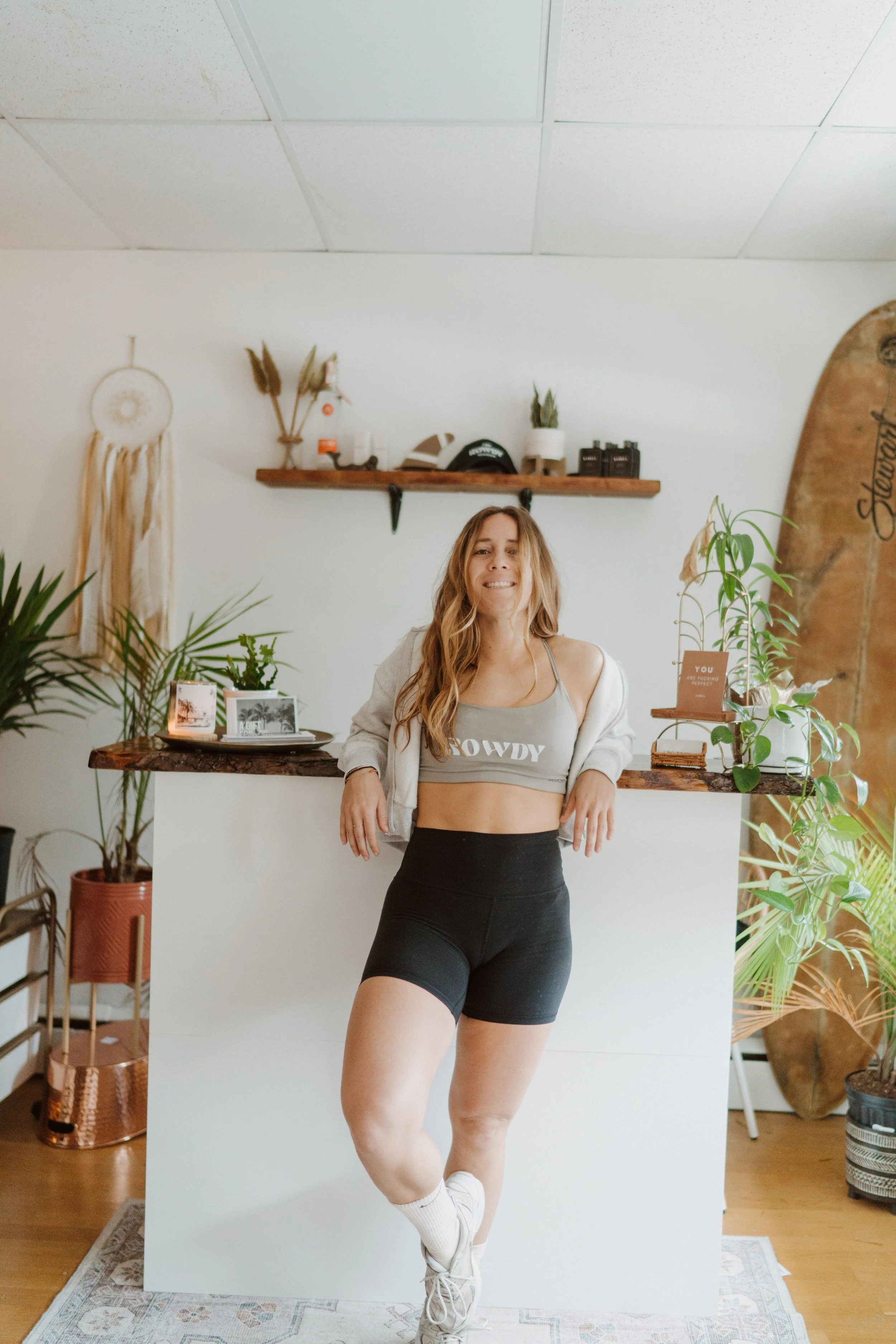 A woman stands in a room with plants, wooden furniture, and a surfboard, wearing black shorts, a gray crop top, and a gray hoodie, with a white wall and shelf behind her.
